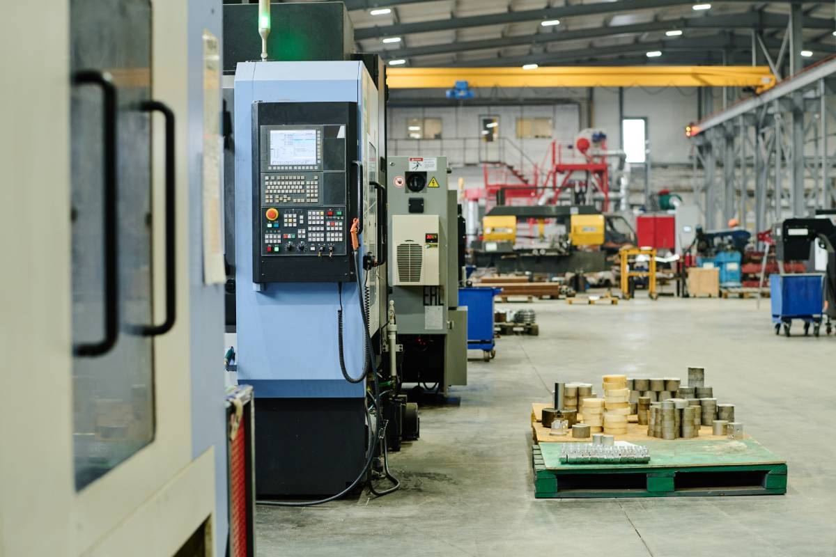 Industrial factory interior with CNC machine, pallets with metal parts, and manufacturing equipment.