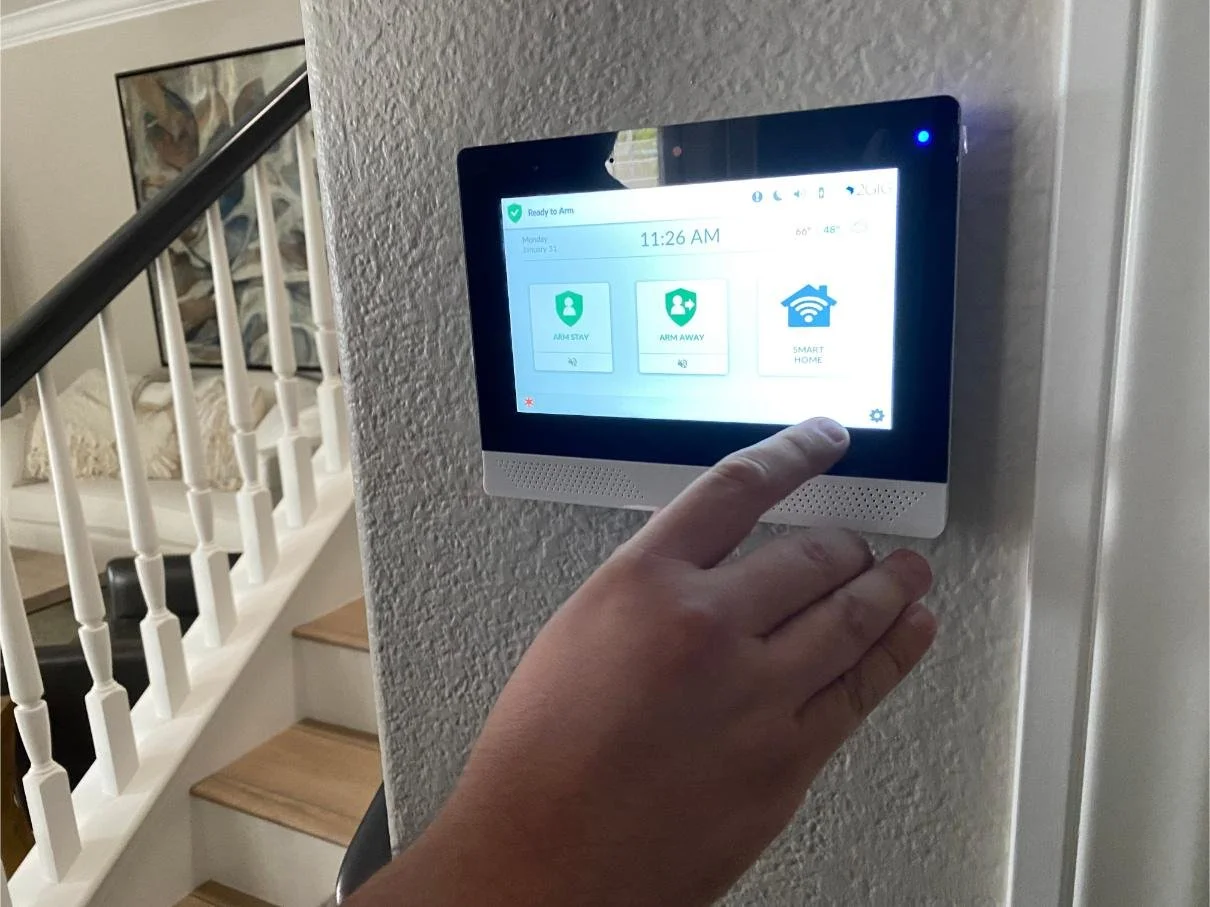 A person is pressing a touchscreen home security control panel mounted on a wall inside a house. The screen displays options for arm stay, arm away, and smart home.