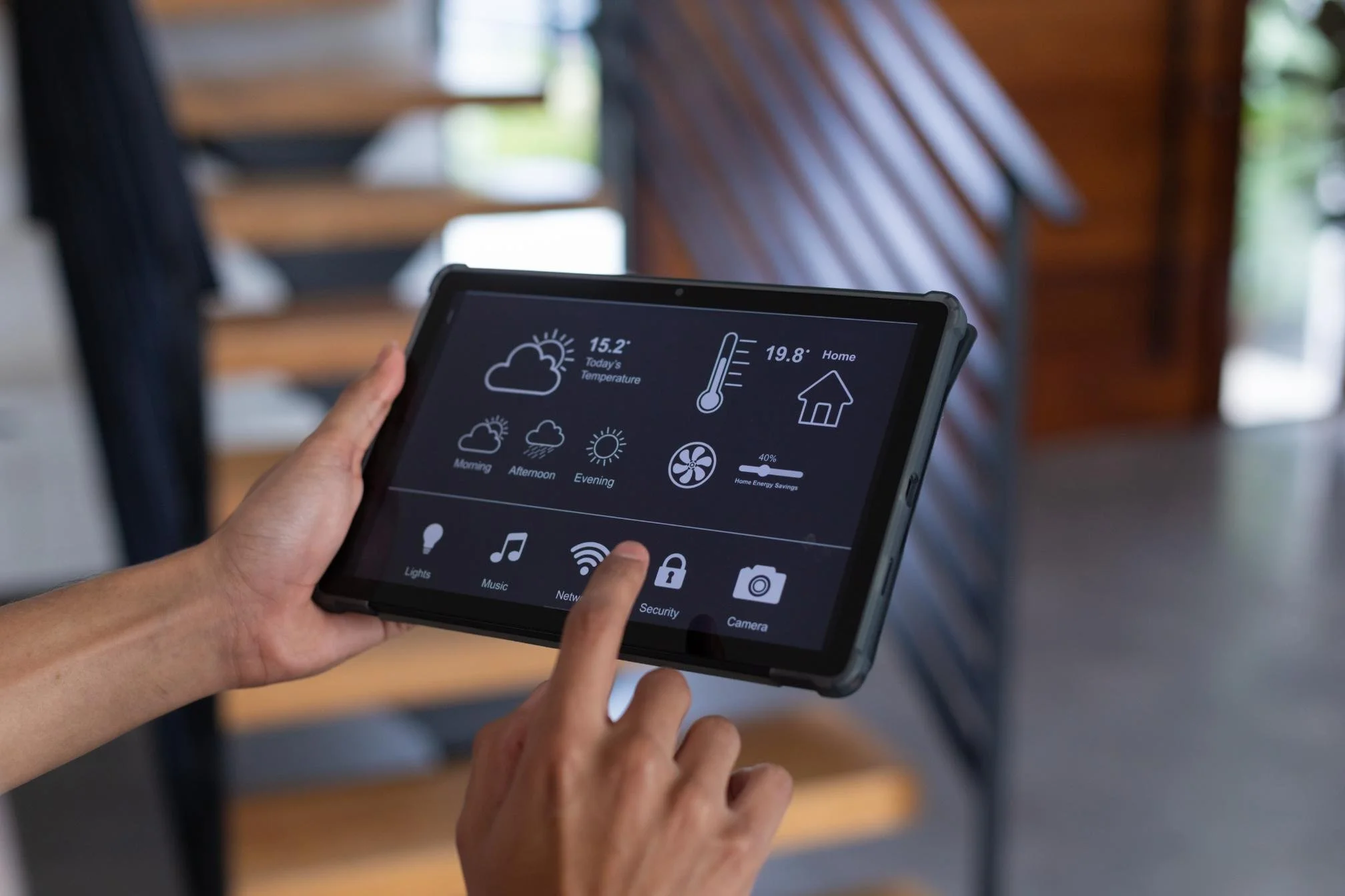 Person holding a tablet with smart home control screen, showing weather, temperature, and icons for lights, music, security, Wi-Fi, camera. Background features a staircase with wooden steps and railing.