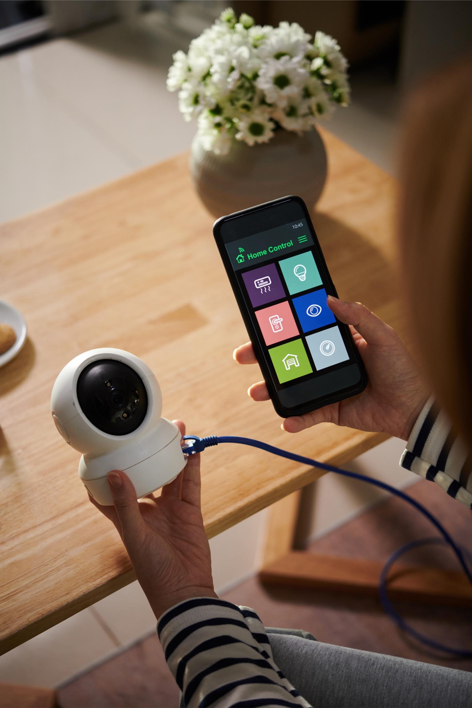 Person holding a small white security camera in one hand and a smartphone in the other, displaying a home control app, with a vase of white flowers on a wooden table in the background.