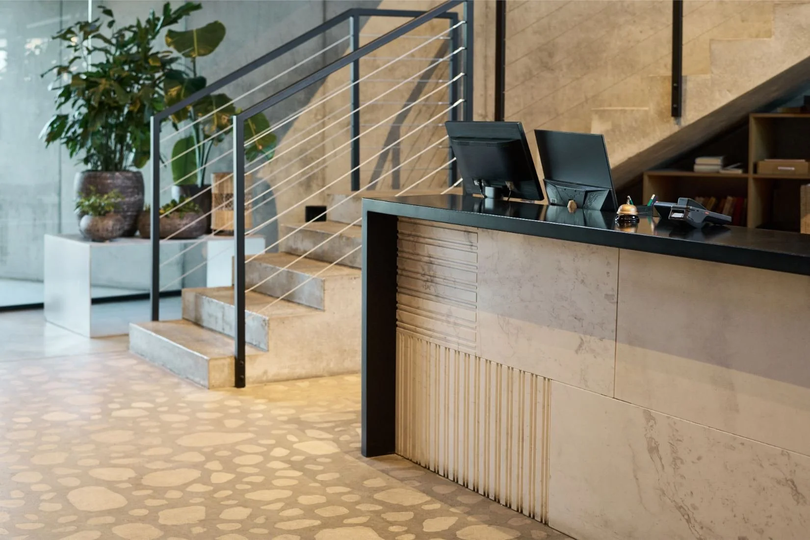 Reception desk with two computer monitors, a phone, and office supplies in a modern lobby area, with stairs, plants, and shelves in the background.