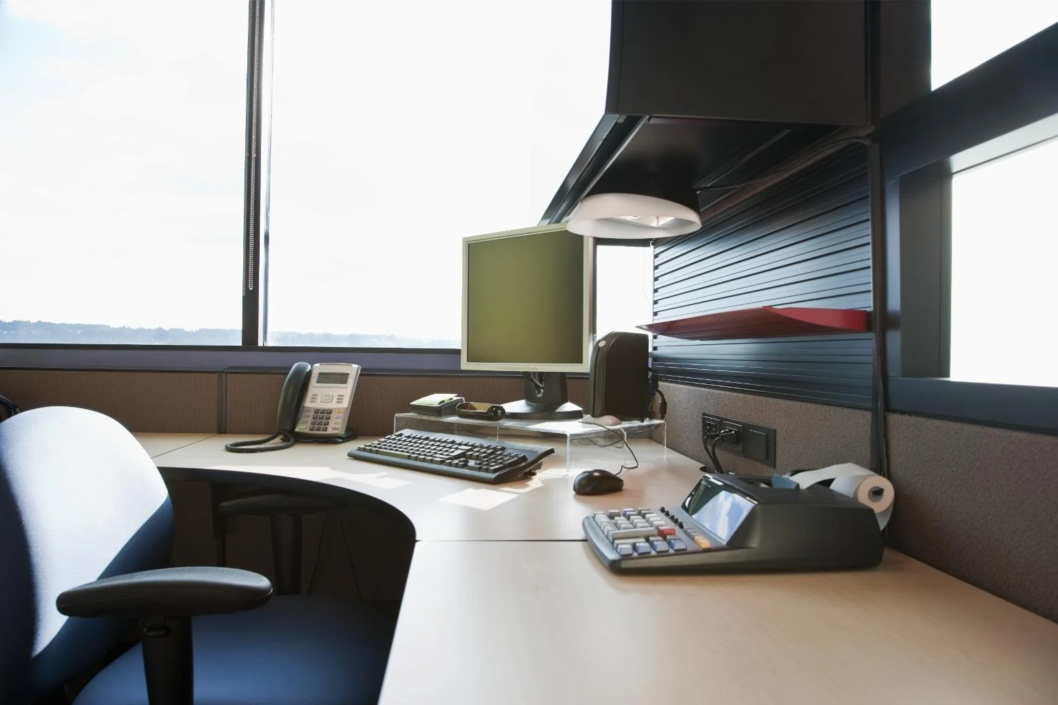 An empty office cubicle with a desk, a computer monitor, keyboard, mouse, telephone, and office supplies, with a window showing a distant landscape in the background.