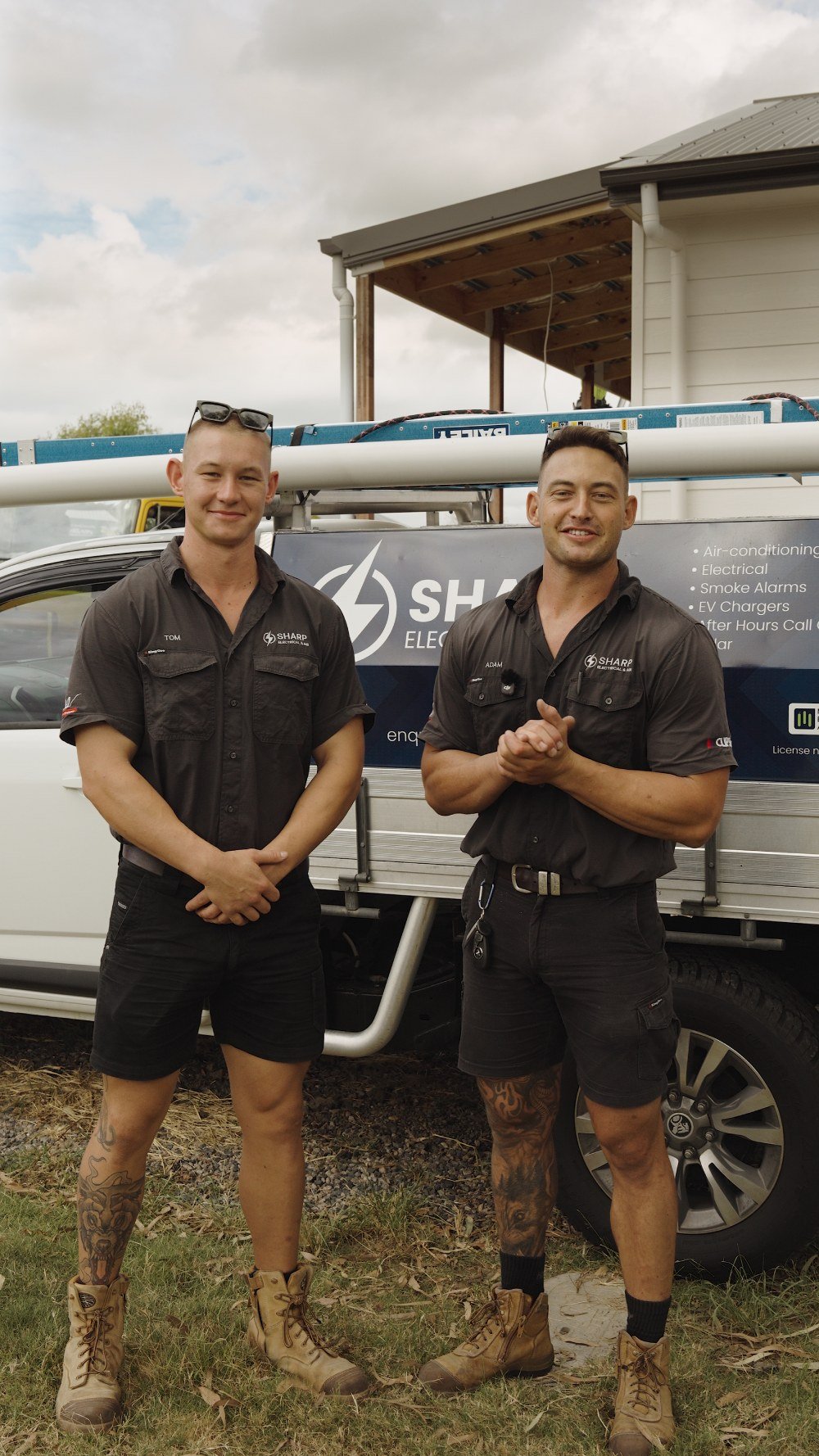 Two men in work uniforms standing in front of a company vehicle with ladders on top, outdoors on a cloudy day.