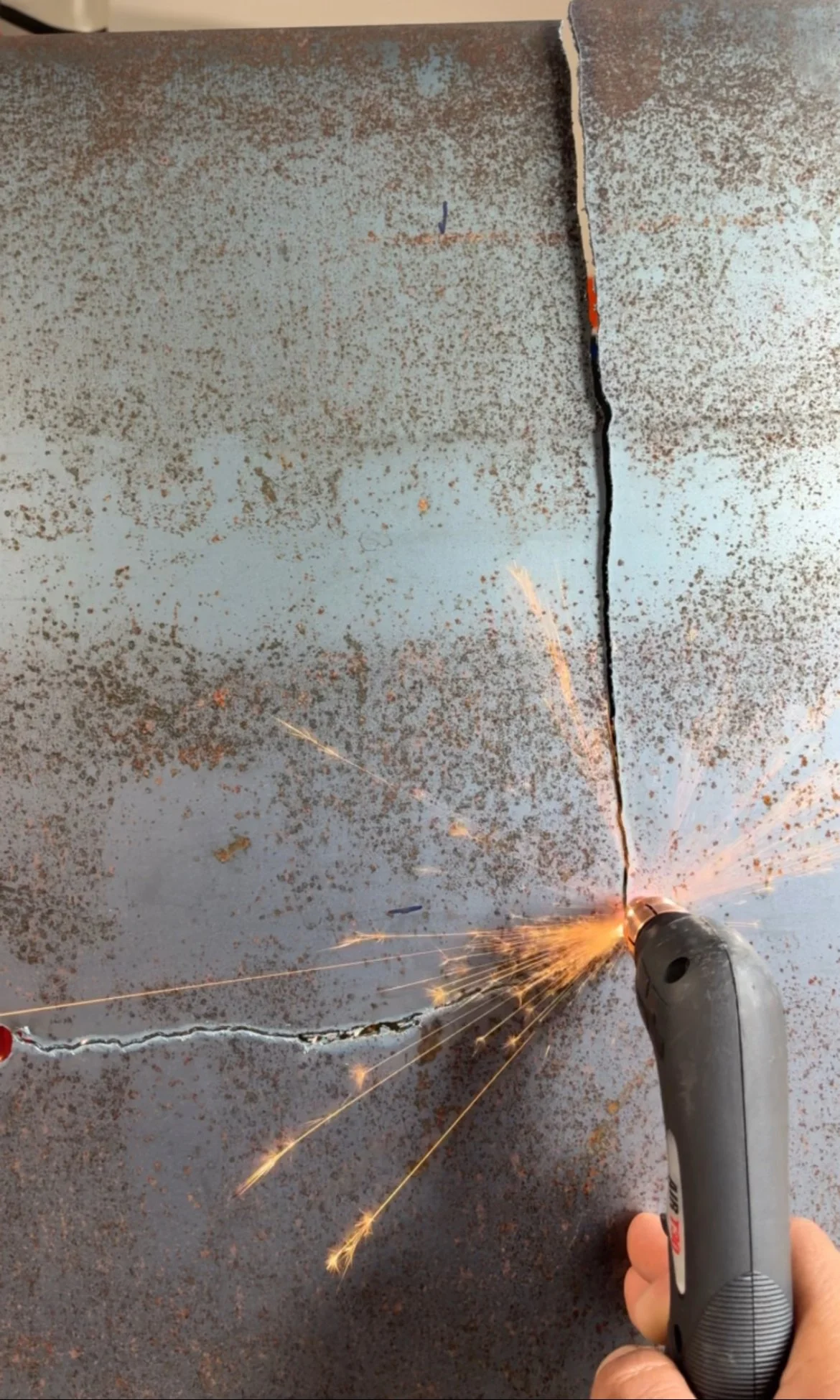 Close-up of a hand holding a plasma cutter creating sparks and cutting through a large, rusted metal surface with visible cracks.