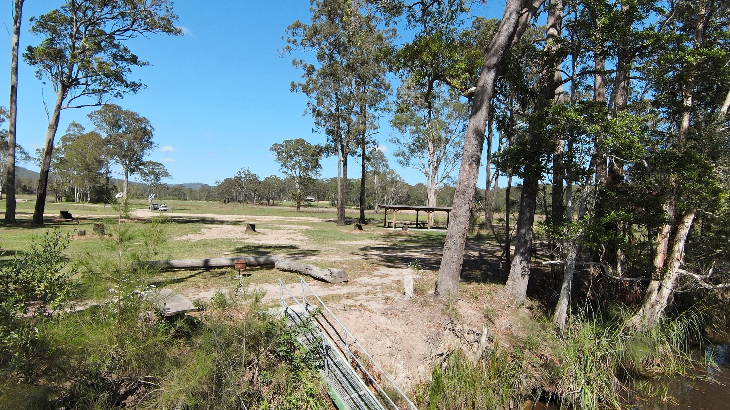 A campground with green grass, trees, a picnic table, a dirt path, a bridge, and cars parked in the distance on a sunny day with blue sky.