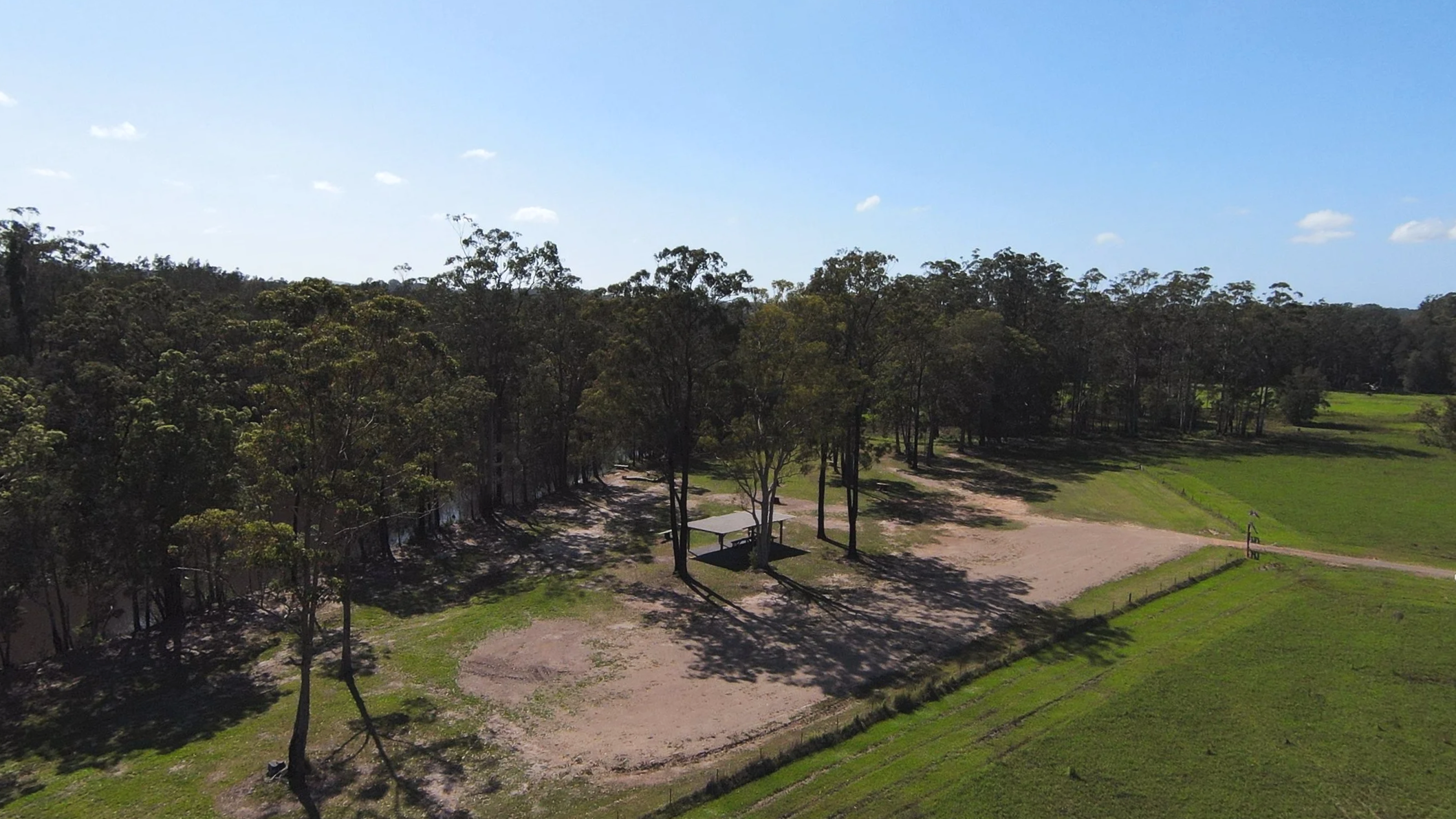 Aerial view of a campground with tall trees, a shaded picnic area with a bench, and open grassy fields under a bright blue sky with a few clouds.