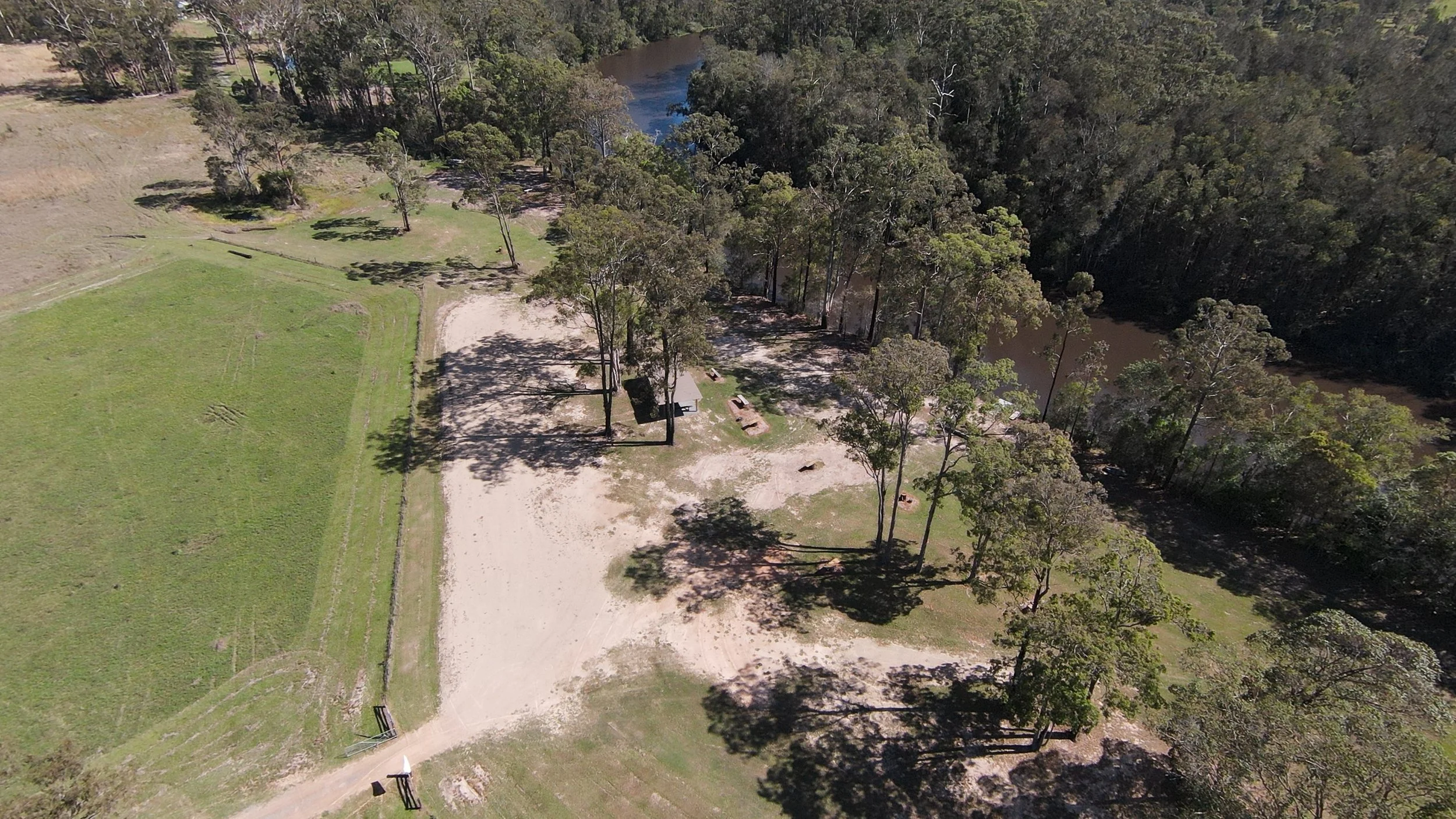 Aerial view of a campground with open grassy areas, trees, a picnic shelter, and nearby a river surrounded by dense woods.