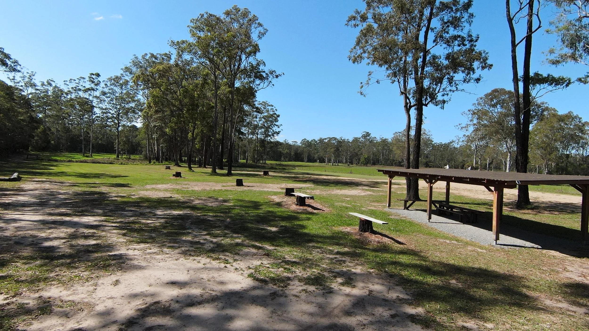 A campground with a grassy open area, trees, and a shaded picnic shelter with a picnic table underneath.