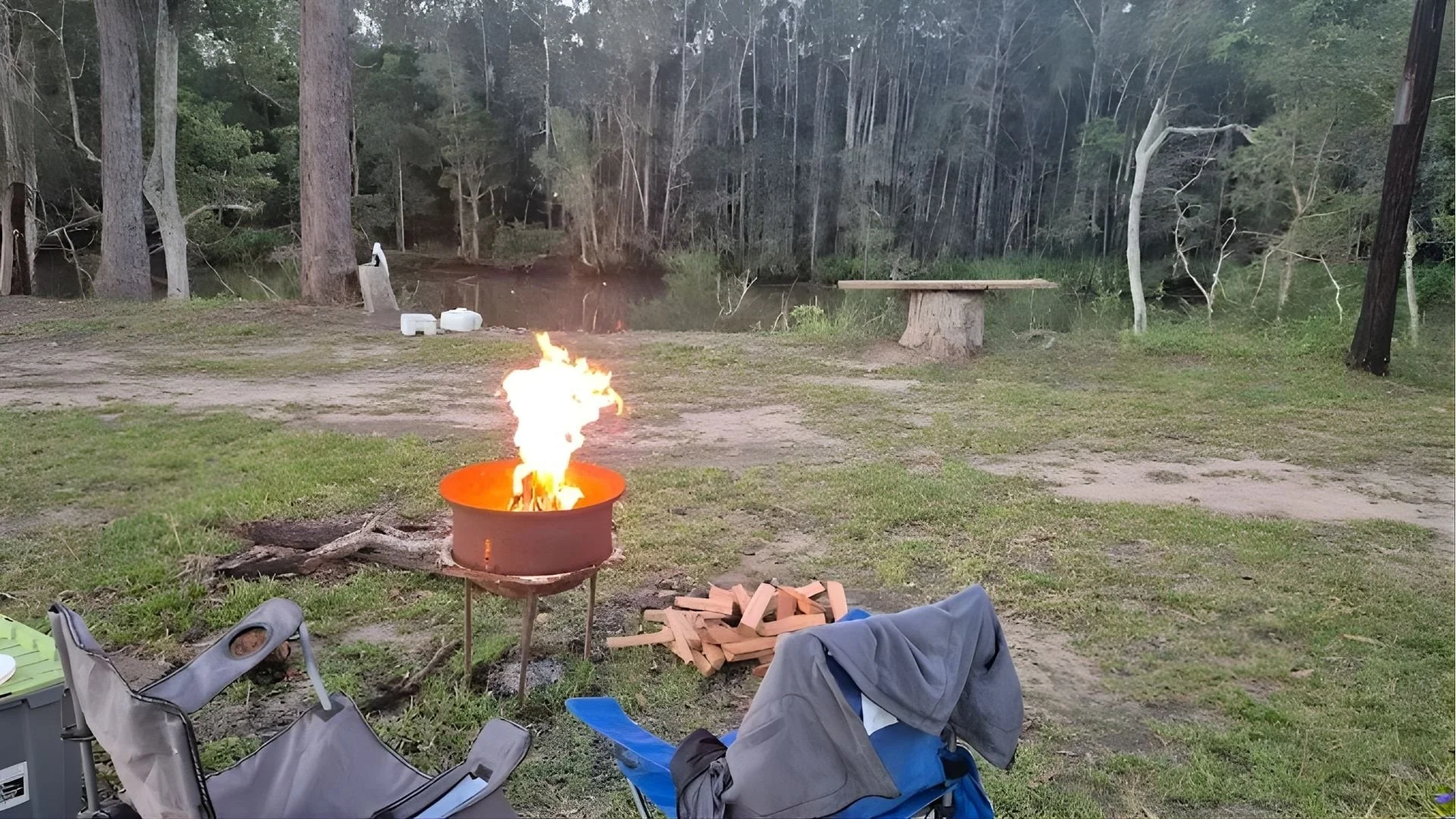Camping scene with a small fire in a metal fire pit, surrounded by camping chairs, and a pile of chopped firewood nearby. In the background, there are trees and a river.