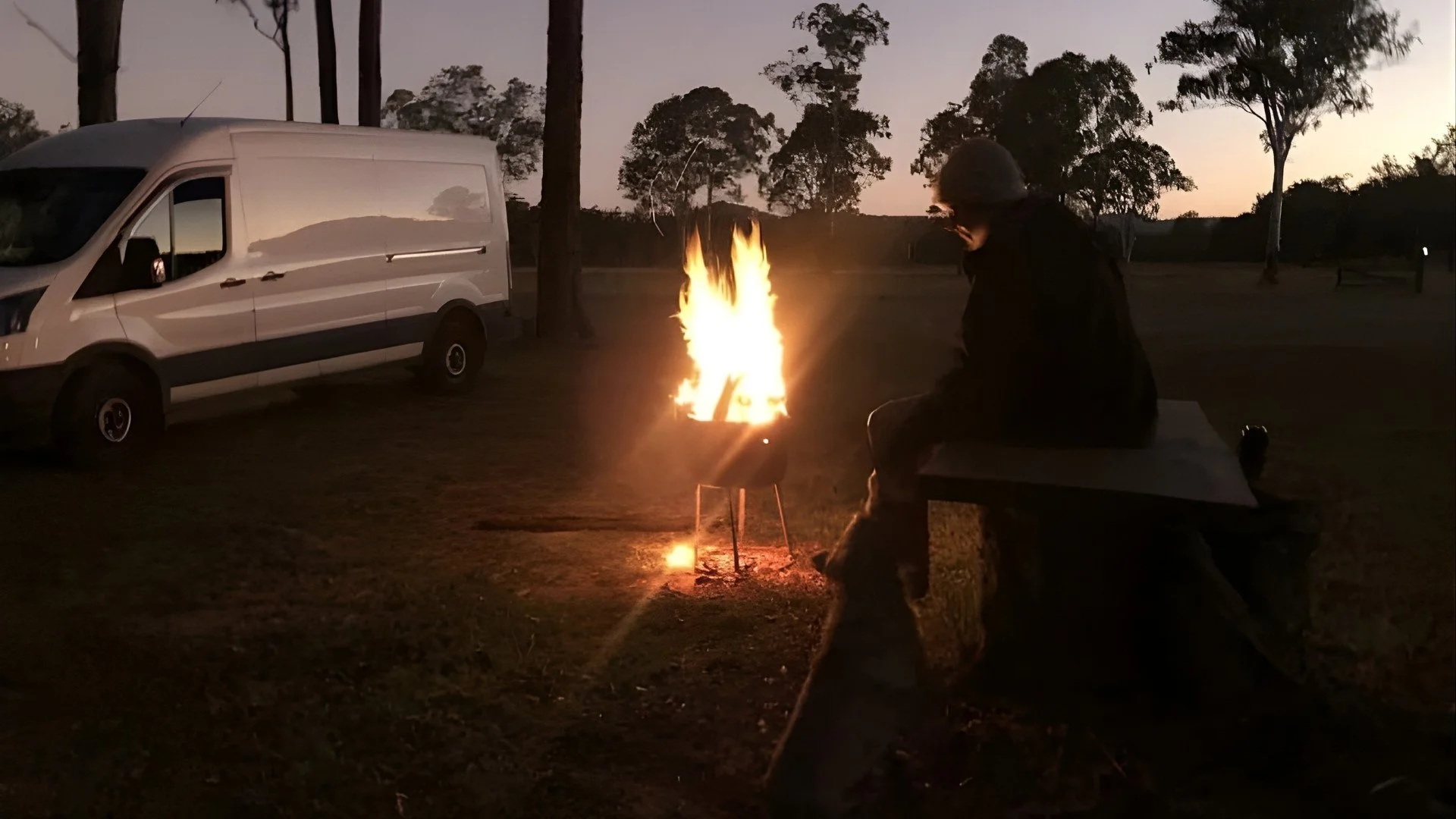 Person sitting on a bench near a campfire in a park during dusk, with a white van parked nearby and trees in the background.