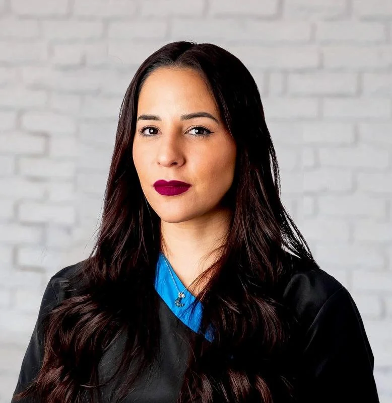 A woman with long, dark hair and dark lipstick, wearing a dark robe with a blue collar, standing in front of a white brick wall.