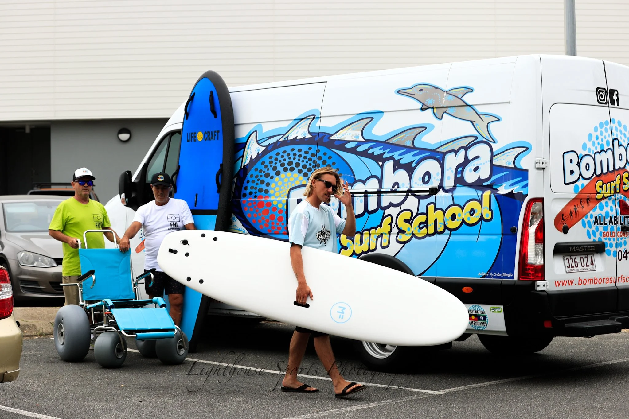 Man with surfboard standing in front of a van with a surf school logo, with two men nearby and a wheelchair, in a parking lot.