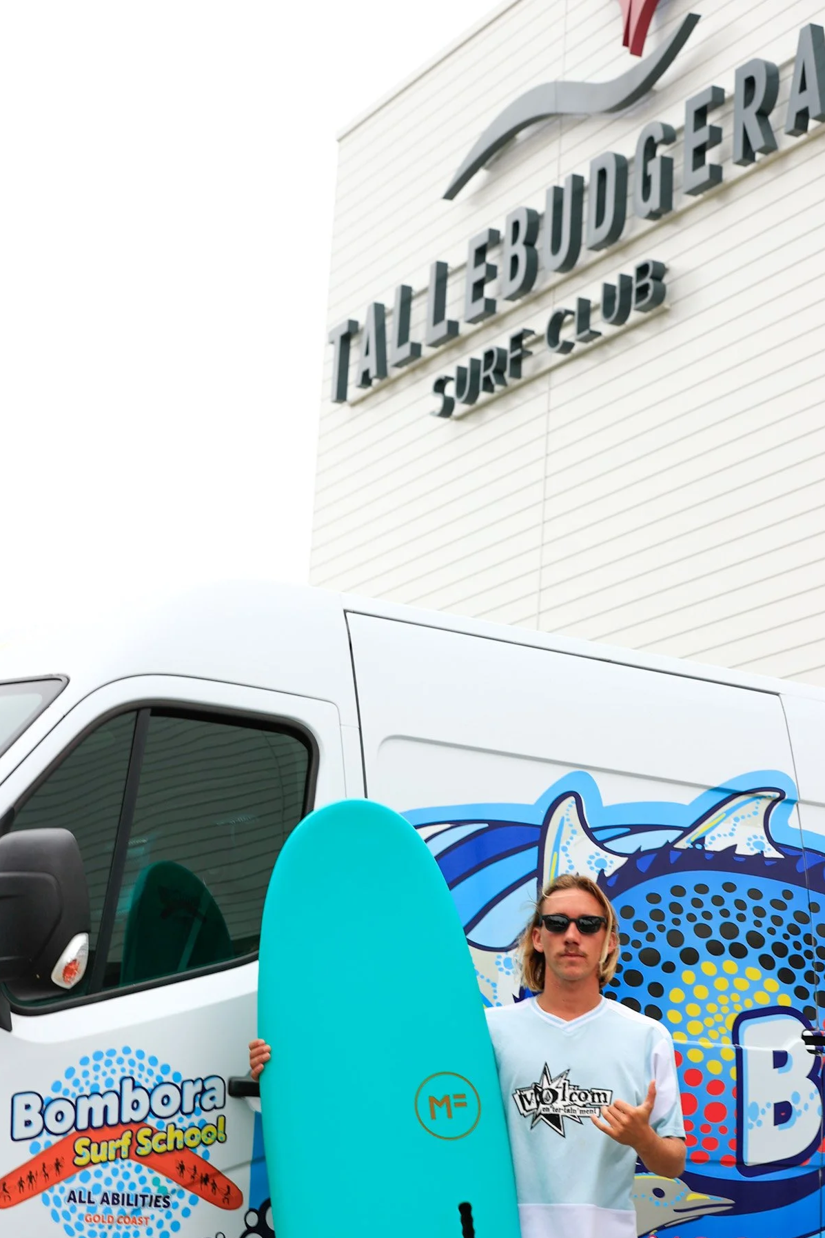 A man holding a blue surfboard in front of a white van with a colorful ocean wave design. The man is wearing sunglasses and a white t-shirt with a logo, standing in front of the Talllebudget Surf Club building.