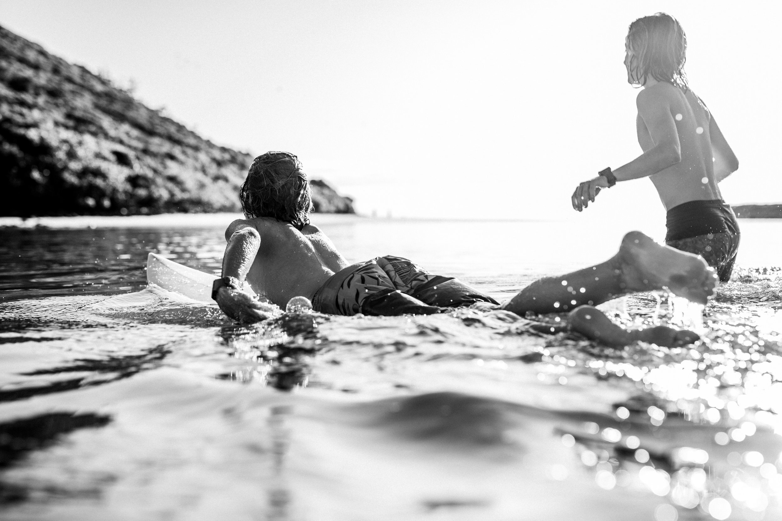 Two children playing in the ocean during sunset, with one lying on their stomach holding a boogie board and the other standing nearby, both wearing swimsuits.
