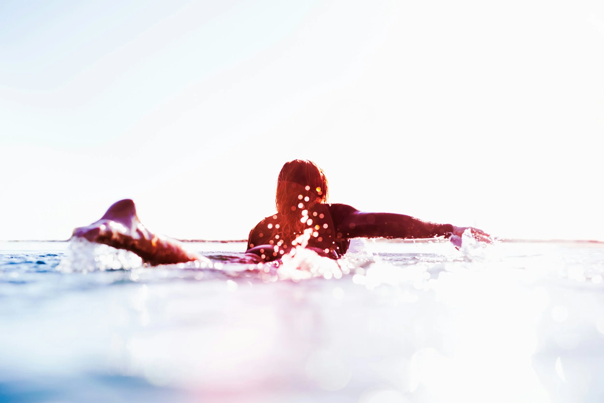 Person surfing in the ocean during sunset, with water splashing around and bright sunlight overhead.