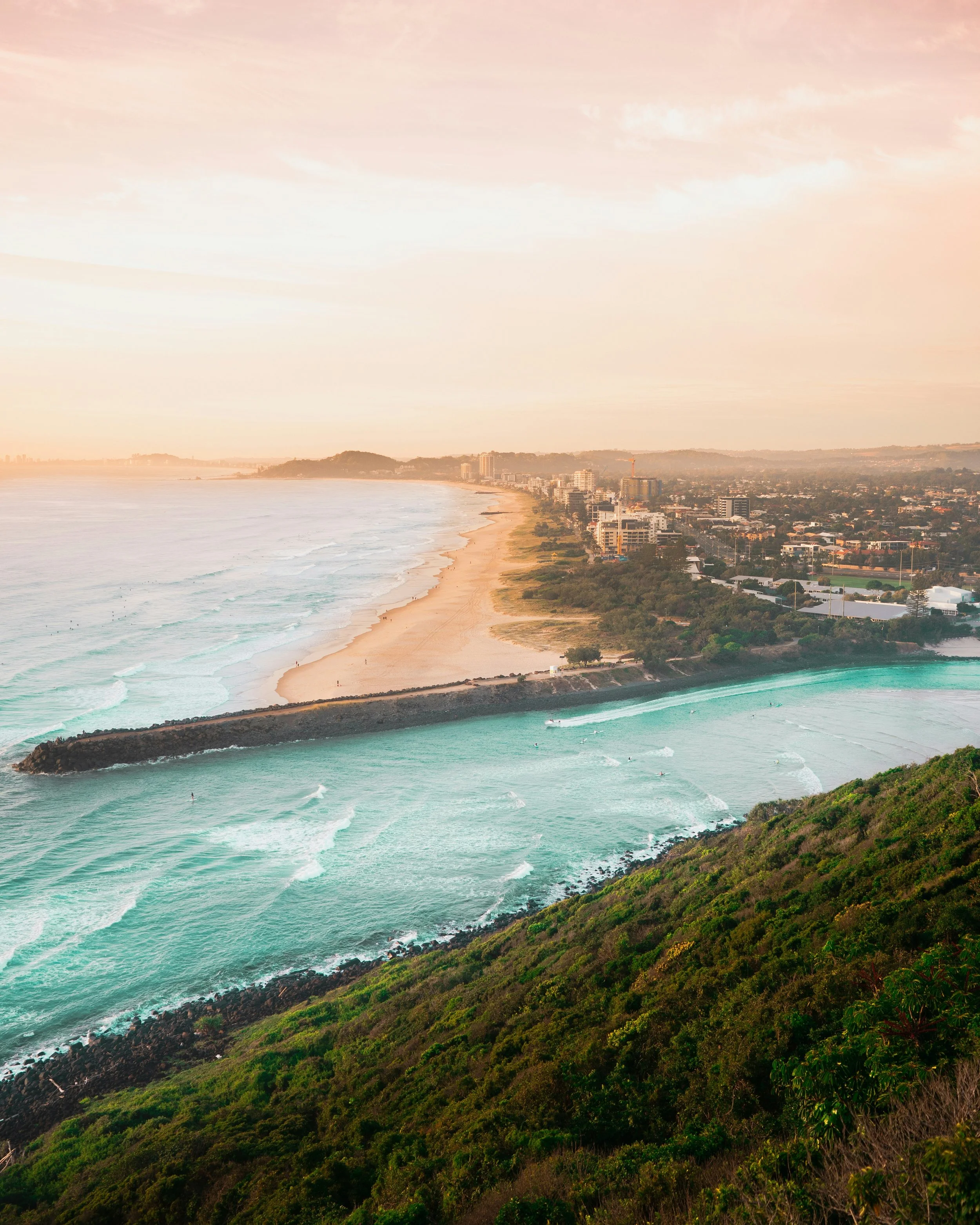 Aerial view of a coastal city at sunset with a sandy beach, ocean waves, and a greenery-covered hillside.