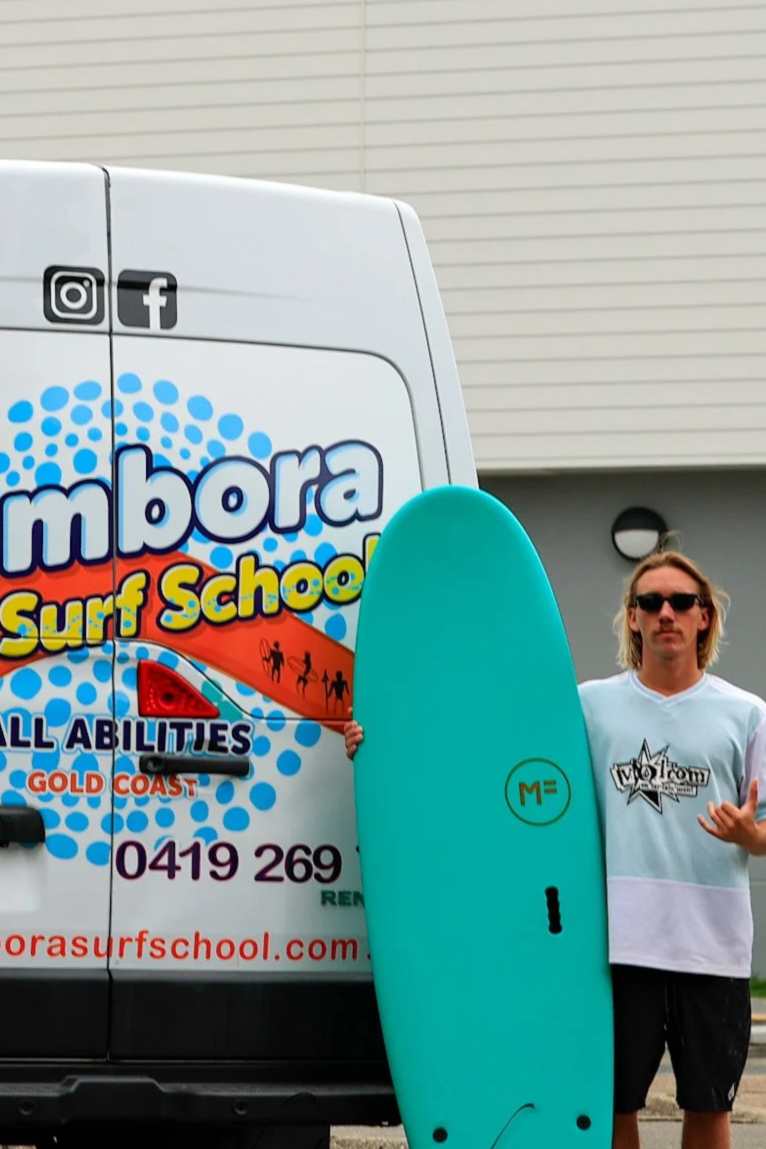 A man holding a blue surfboard stands in front of a white van with colorful surf school logo and social media icons.