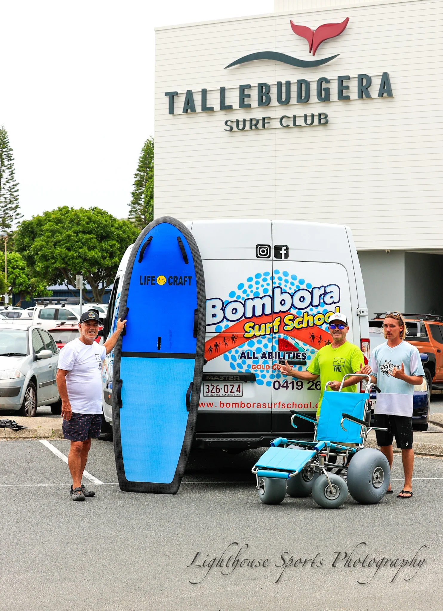 Three men standing in a parking lot with surf equipment, including a large paddleboard, a wheelchair, and a van with the logo for Bombora Surf School in front of a Tallebudgera Surf Club building.