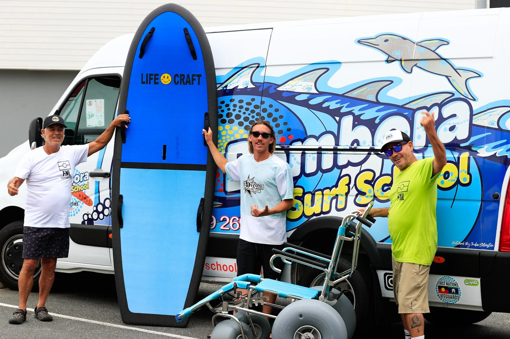Three men standing in front of a colorful van with a surf school logo, holding a blue stand-up paddleboard and a wheelchair. One man is wearing a white t-shirt and shorts, the second man is in a white surf jersey, and the third man is in a neon yellow t-shirt and beige shorts. They are smiling and celebrating.