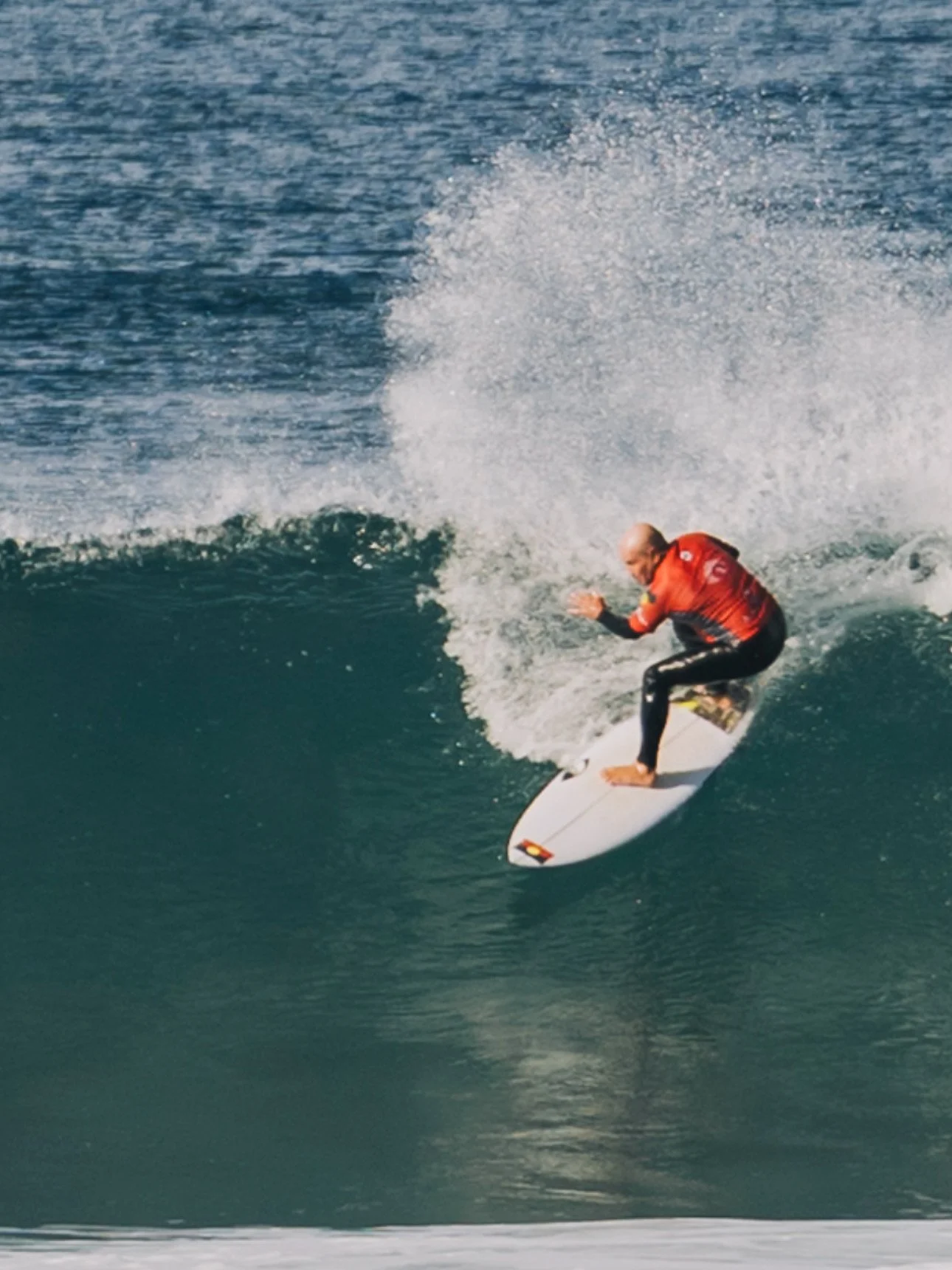 A surfer riding a wave on a sunny day, wearing a red rash guard and black wetsuit, balancing on a white surfboard.