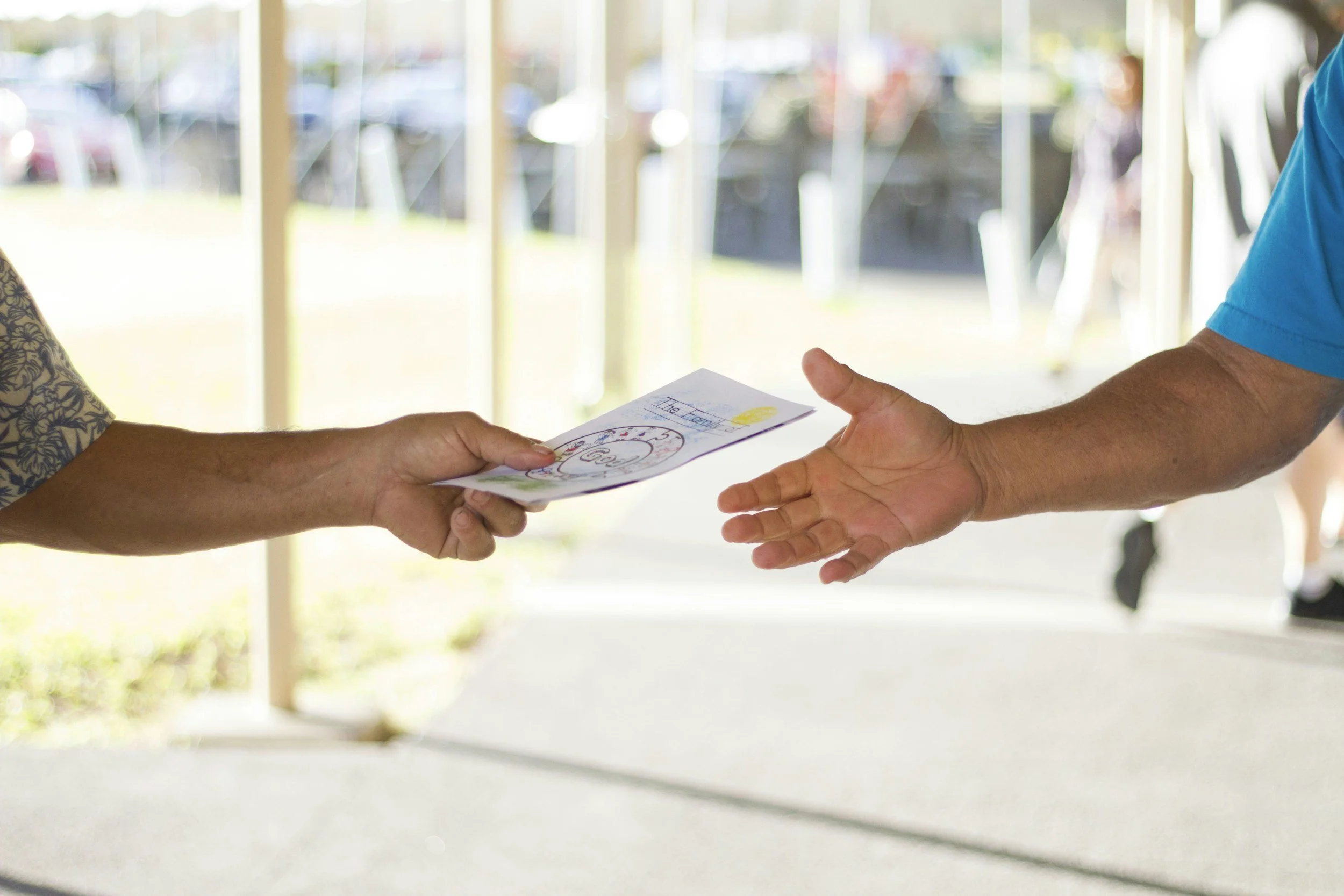 Person in a patterned shirt hands a colorful flyer or brochure to a man in a blue shirt, outdoor setting with blurred background.