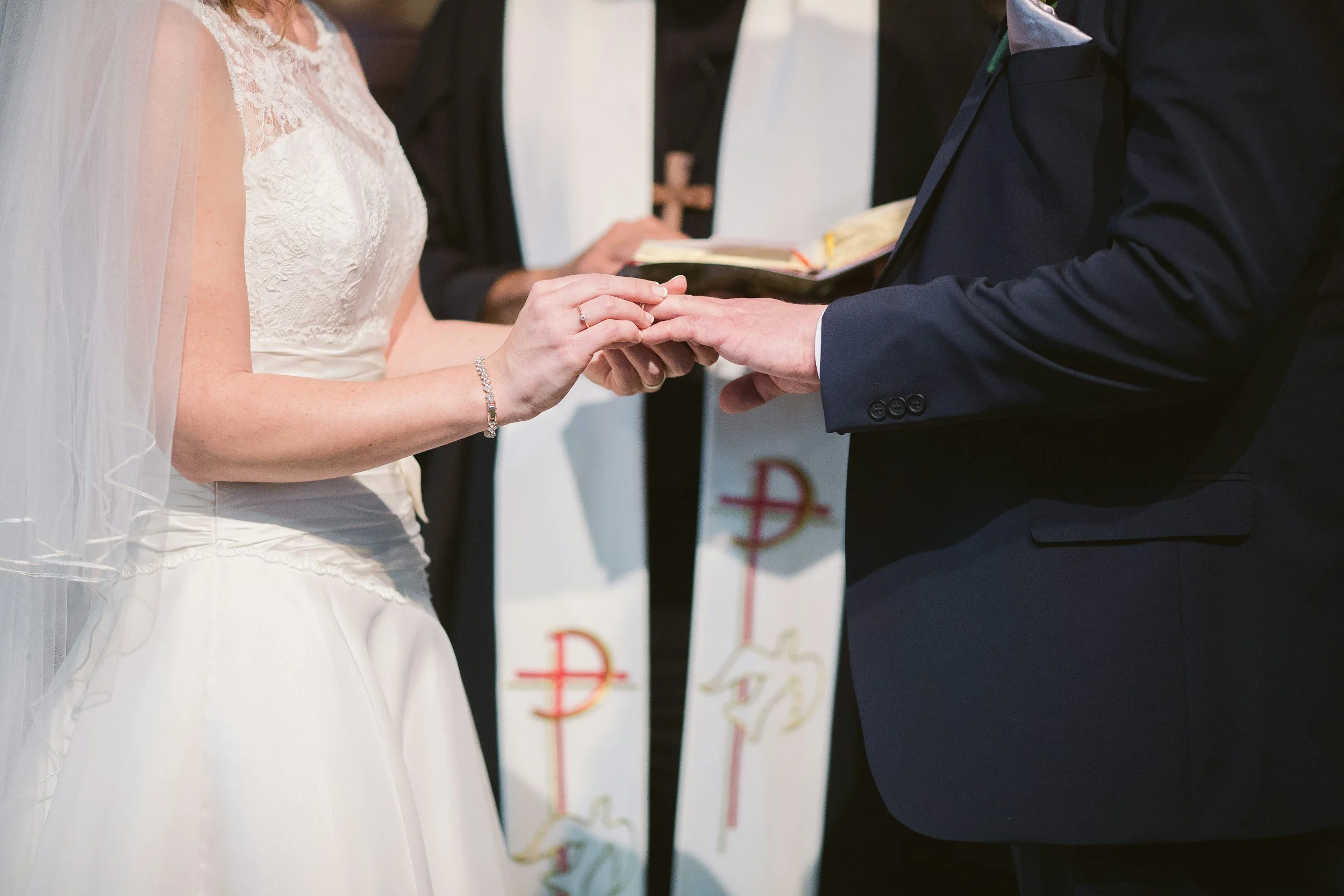 A bride and groom exchanging wedding rings during a wedding ceremony, with an officiant holding a book in the background.