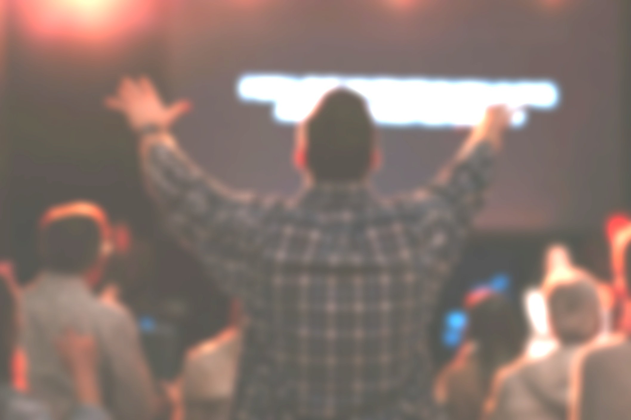 A person with short hair and a dark checkered shirt standing with arms outstretched in front of a blurred audience at a presentation or event, with a glowing digital screen behind them.