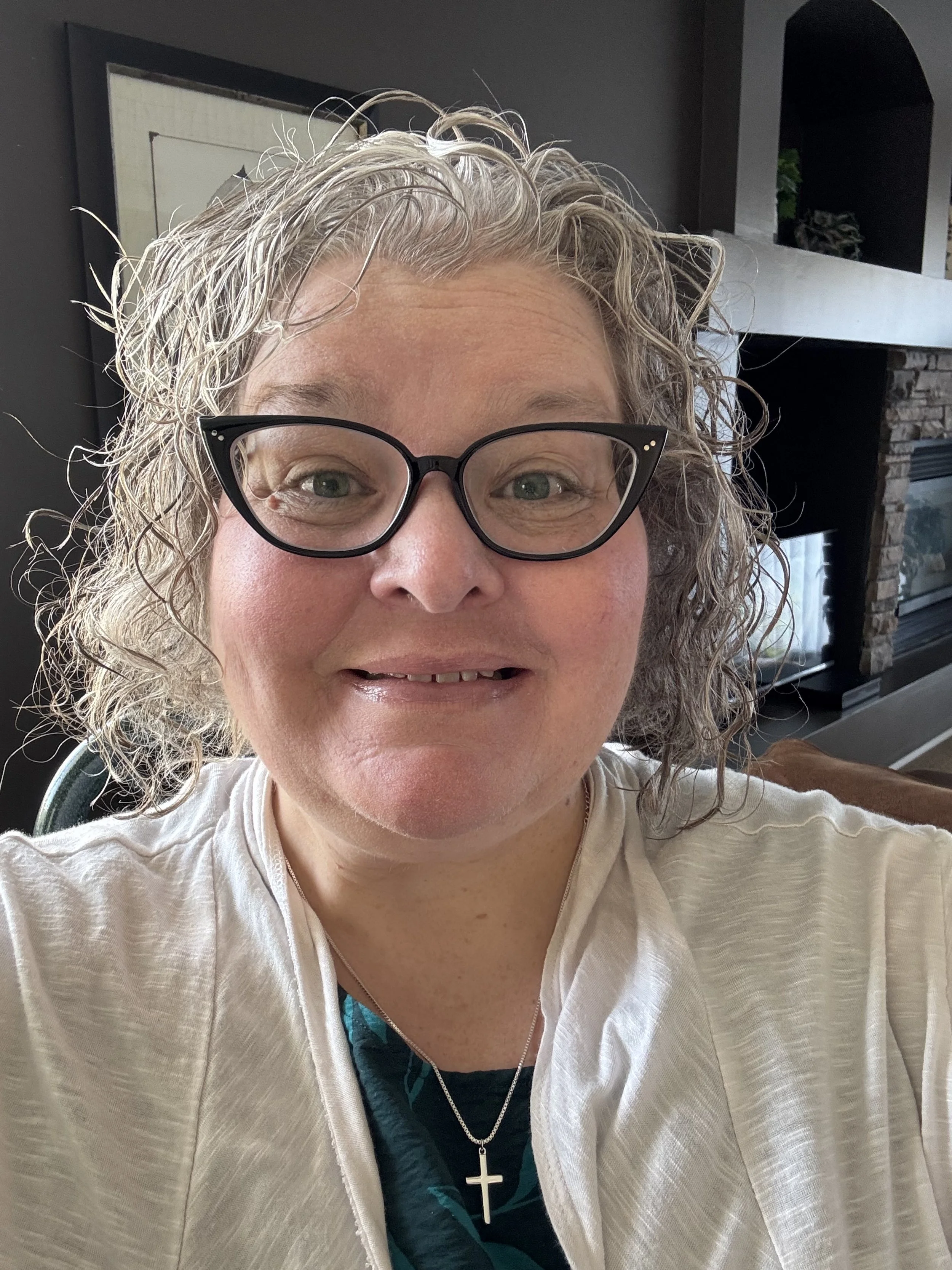 A woman with curly gray hair, wearing glasses, a white shirt, and a cross necklace, smiling for a selfie indoors with a fireplace and television in the background.