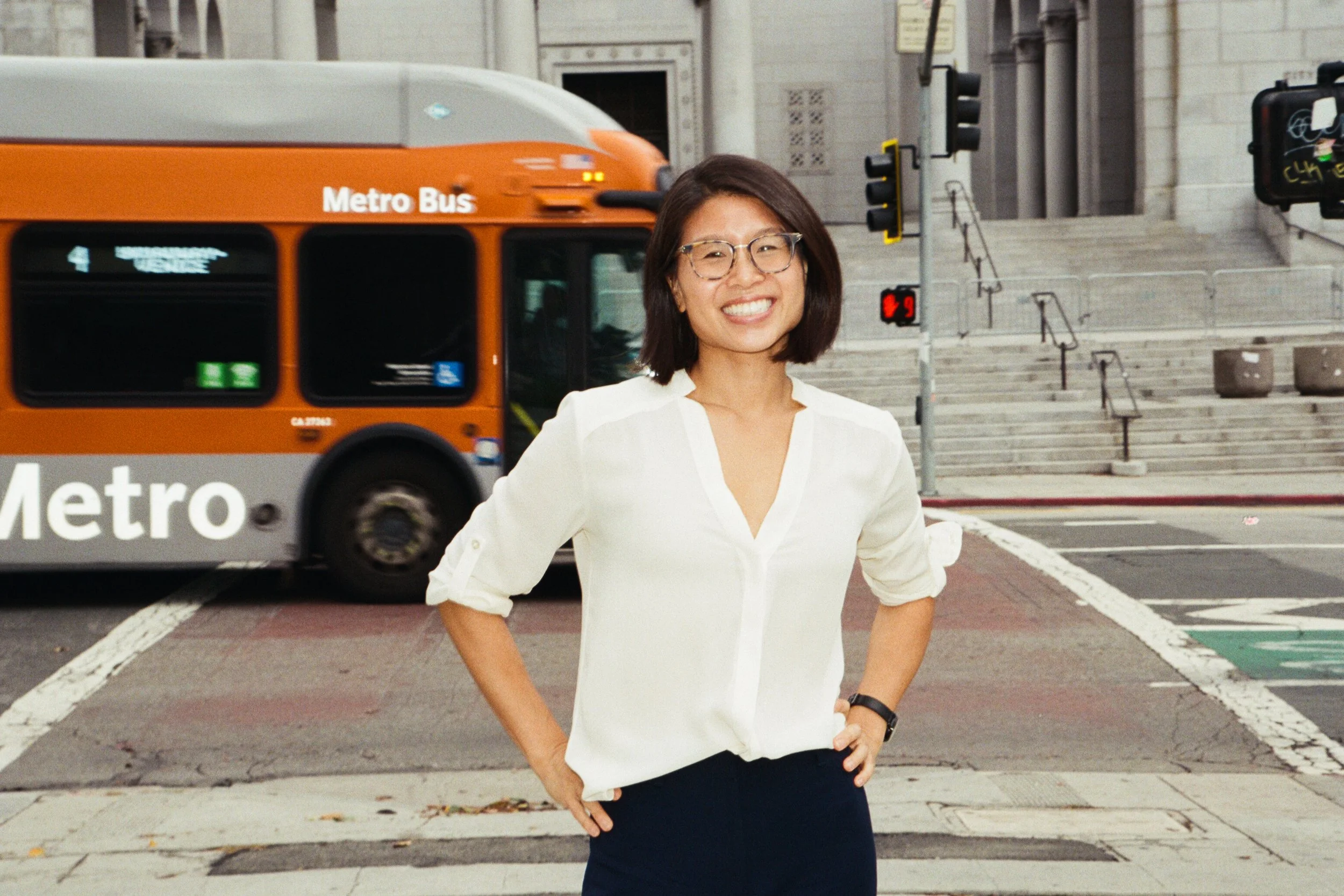 A woman with glasses and shoulder-length dark hair wearing a cream-colored blouse and dark pants, smiling with hands on her hips, standing at a city street corner with a bus and steps in the background.