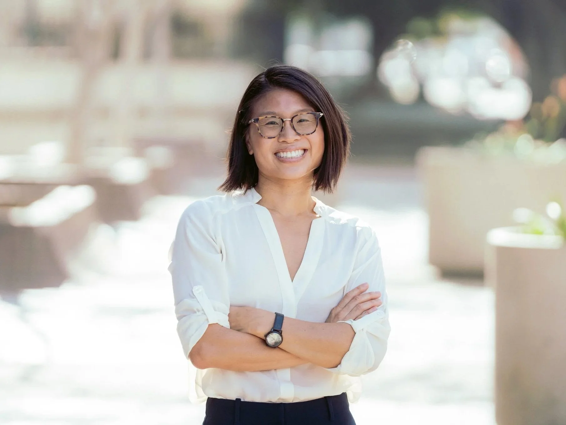 A smiling woman with glasses, wearing a white shirt and a wristwatch, standing outdoors with arms crossed.