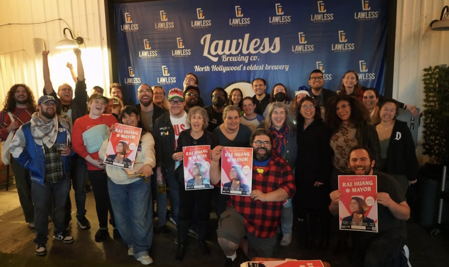 Group of people smiling and posing for a photo in front of a blue backdrop with 'Lawless Brewing Co.' branding, some holding campaign posters for Rae Huang for Mayor, inside a well-lit room.