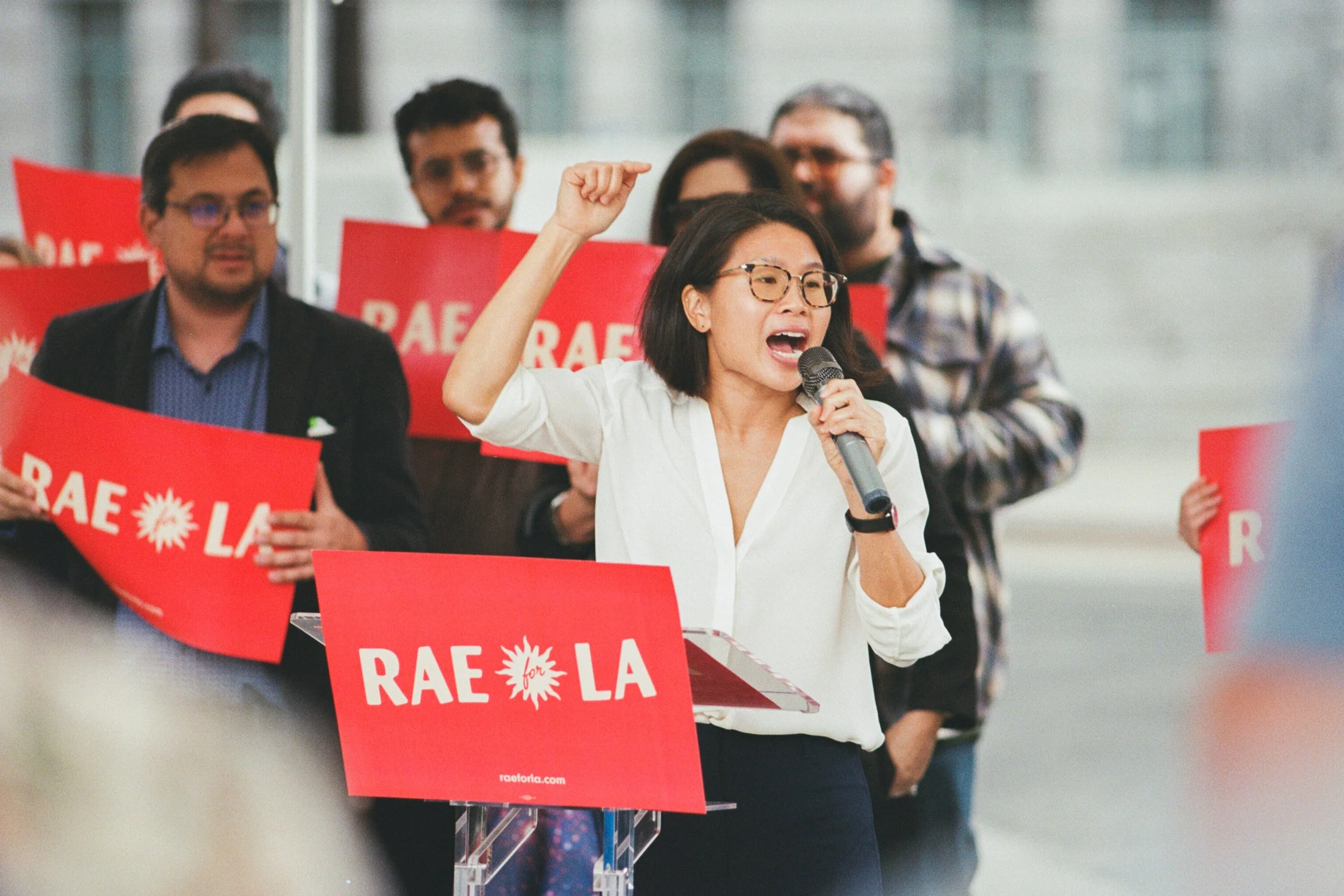 A woman with glasses and a white shirt speaking into a microphone at a rally or protest, with people holding red signs that say 'RAE for LA' in the background.