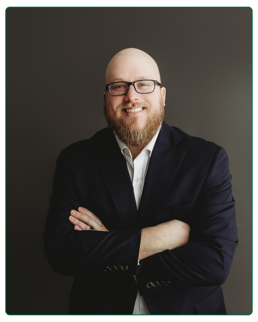 Portrait of a smiling man with glasses, a beard, a white shirt, and a dark blazer against a gray background.