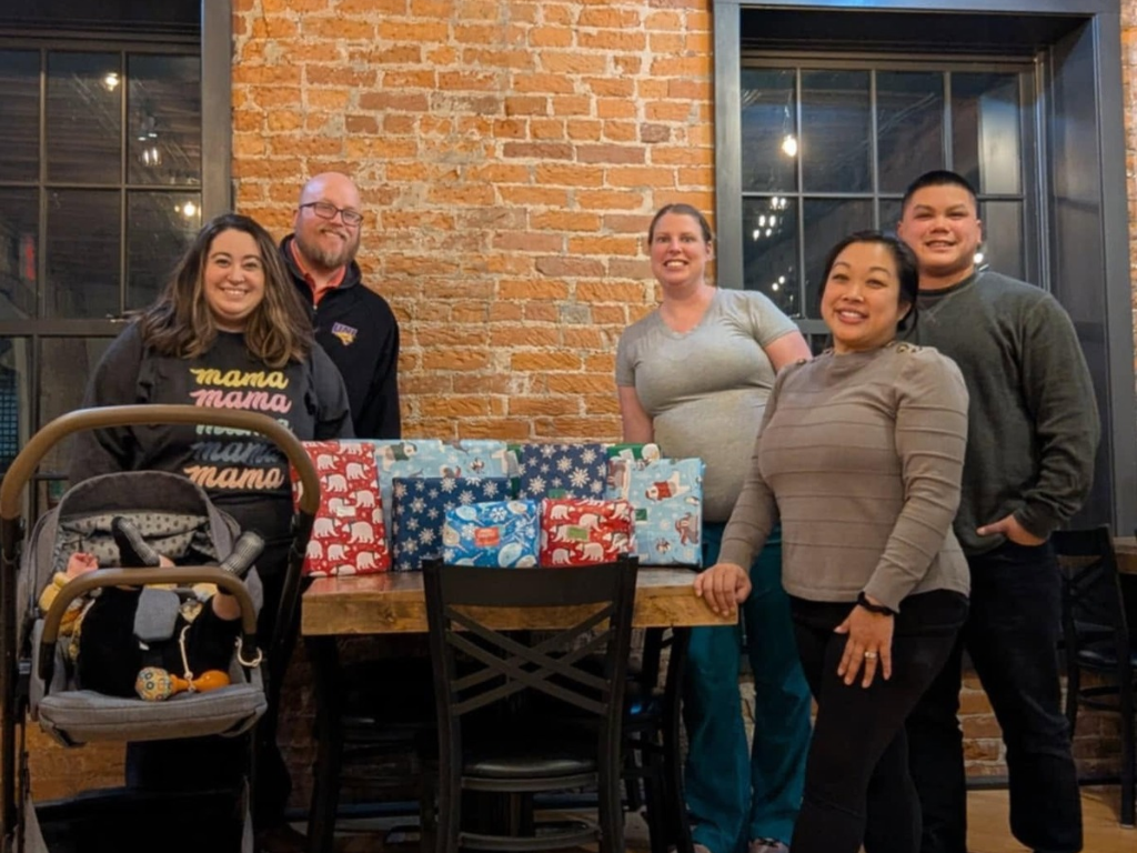 Group of five adults standing behind a table with wrapped Christmas presents in a restaurant or cafe with brick walls and large windows.