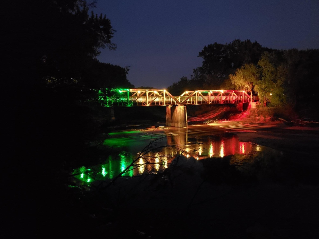 A bridge illuminated with green and red lights reflects on a river at night, surrounded by trees.