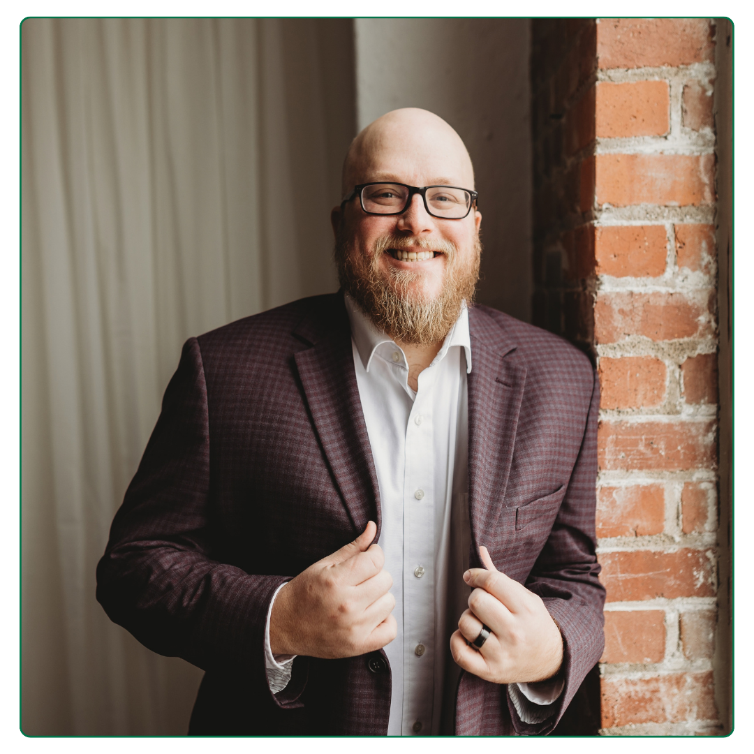 A smiling man with glasses, a beard, and a bald head, dressed in a white shirt and a checkered blazer, standing next to an exposed brick wall inside a room with curtains.