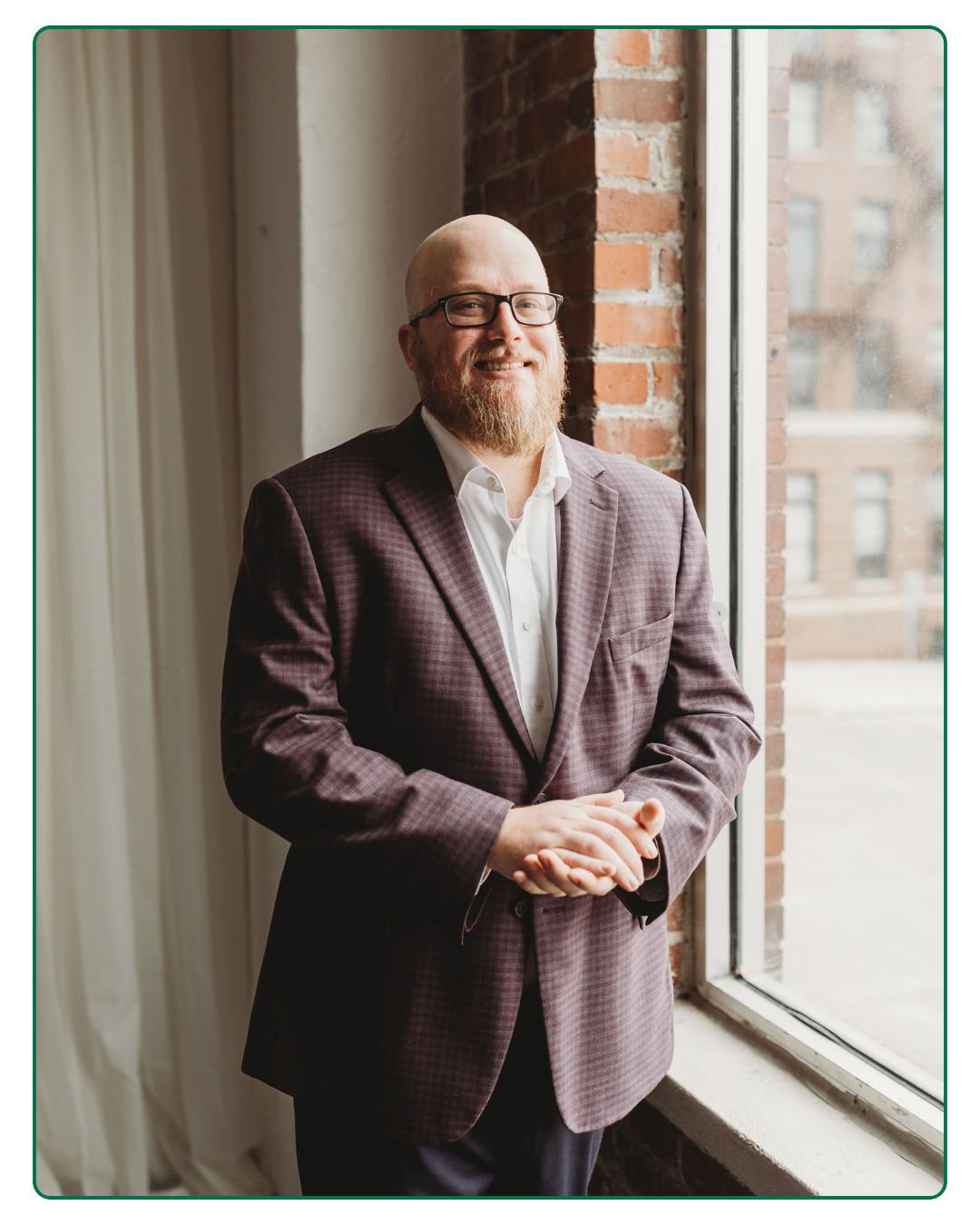 A man with glasses and a beard in a checkered blazer standing by a large window with brick walls visible outside.