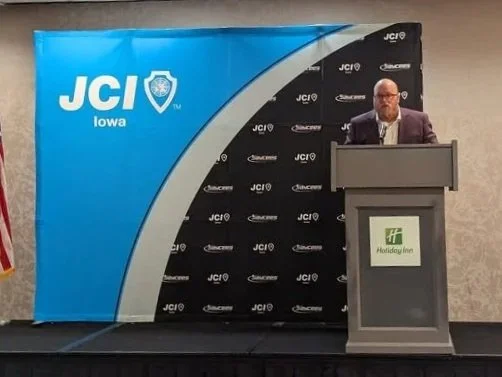 Man speaking at a podium during a JCI Iowa event, with a blue and black backdrop featuring the JCI logo and Holiday Inn logo.