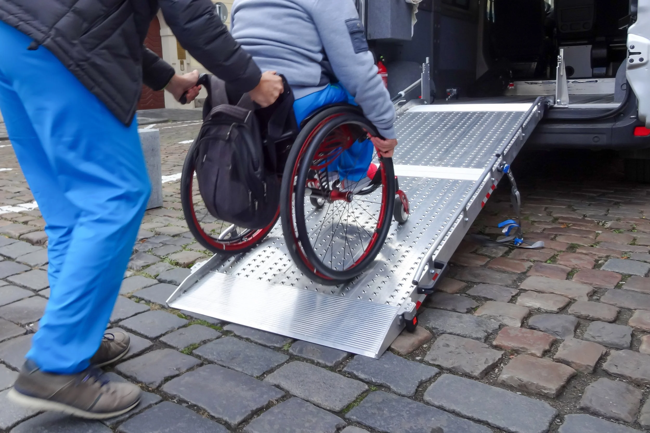 Person in blue wheelchair being assisted onto a vehicle with a ramp by another person.