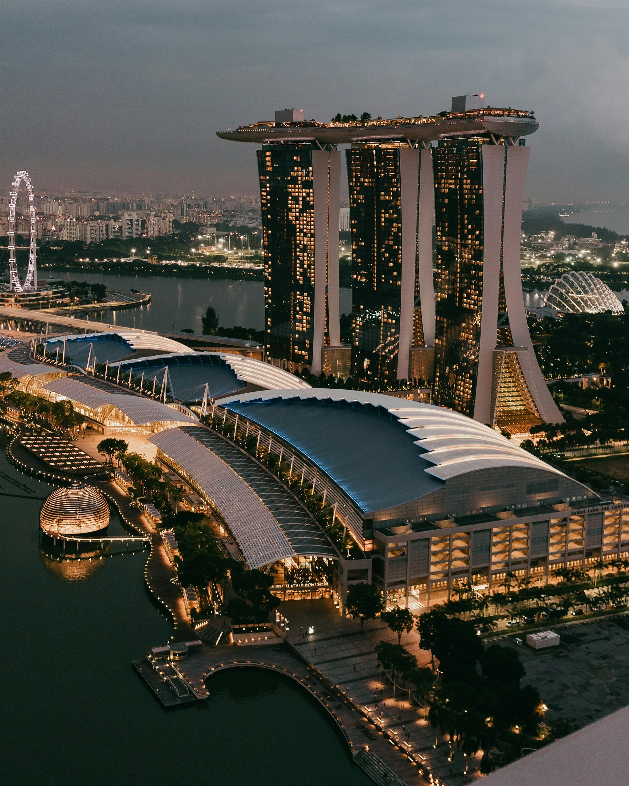 Nighttime aerial view of Marina Bay Sands hotel with its three towers and a boat-shaped rooftop, with surrounding modern structures and city skyline.