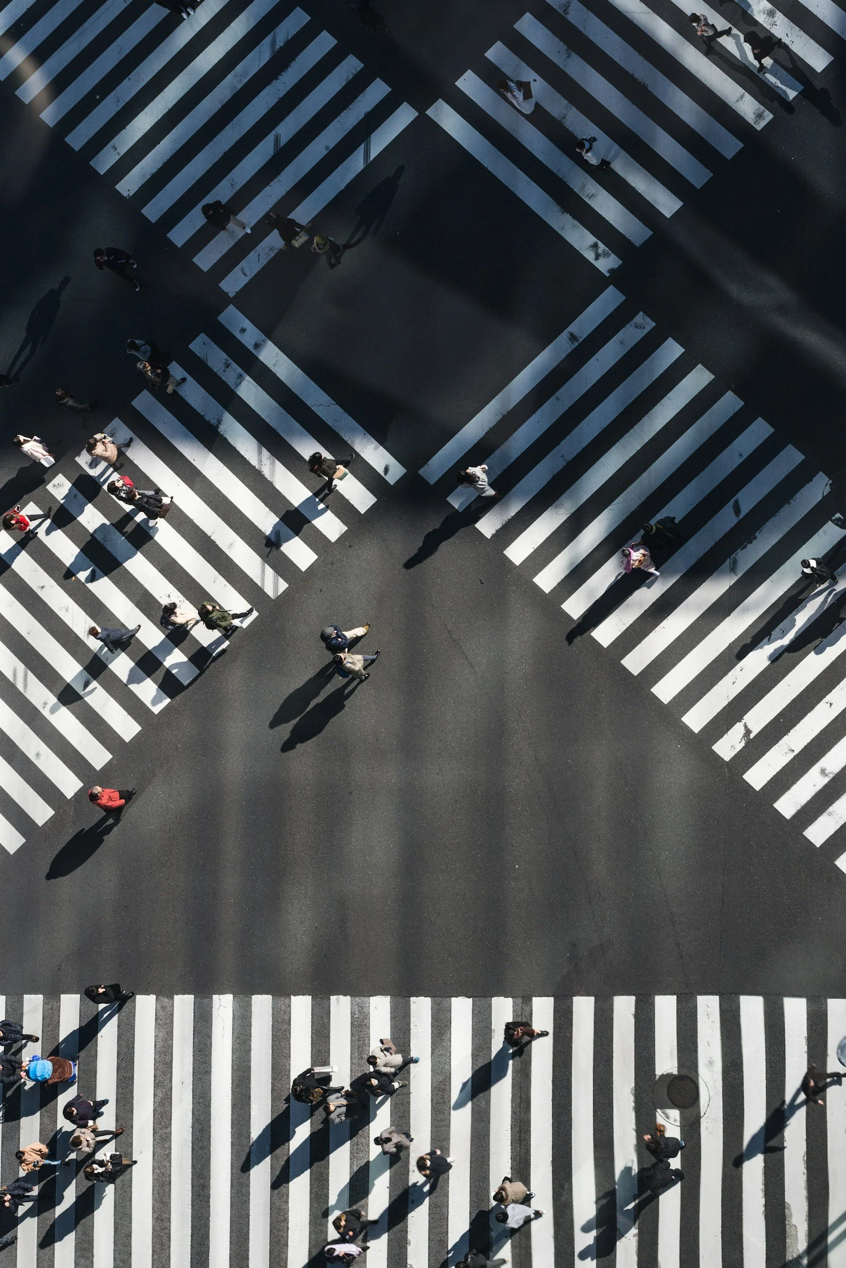 Bird's eye view of pedestrians crossing streets at a busy intersection with marked crosswalks, shadows cast by the people and crosswalk markings.