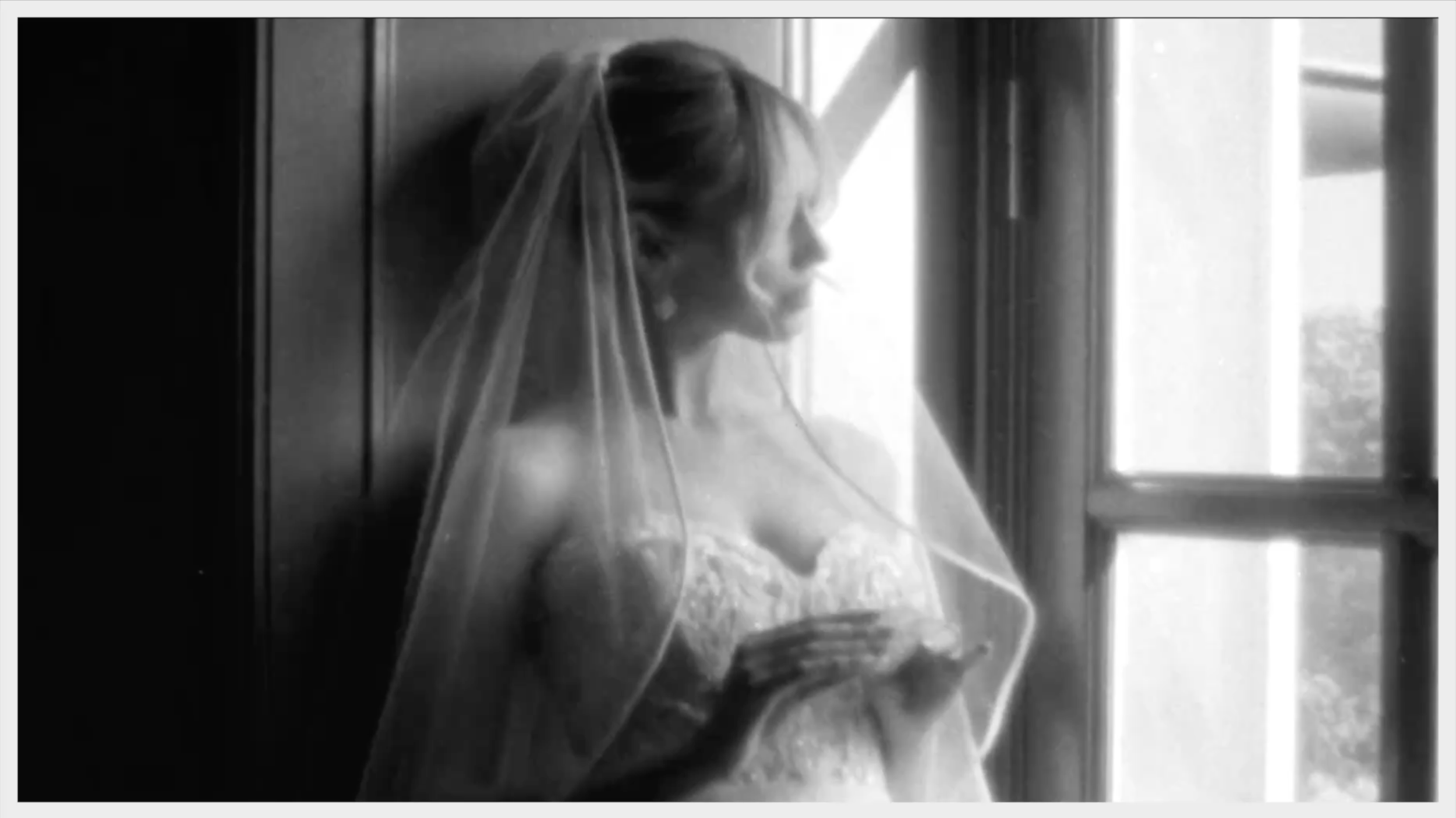 Black-and-white photo of a woman in a wedding dress with a veil, standing near a window, looking at her phone.