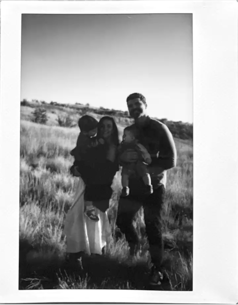 A black and white photo of a family of four outdoors in a grassy field with hills in the background. The mother and father are holding each of their children, and everyone is smiling.