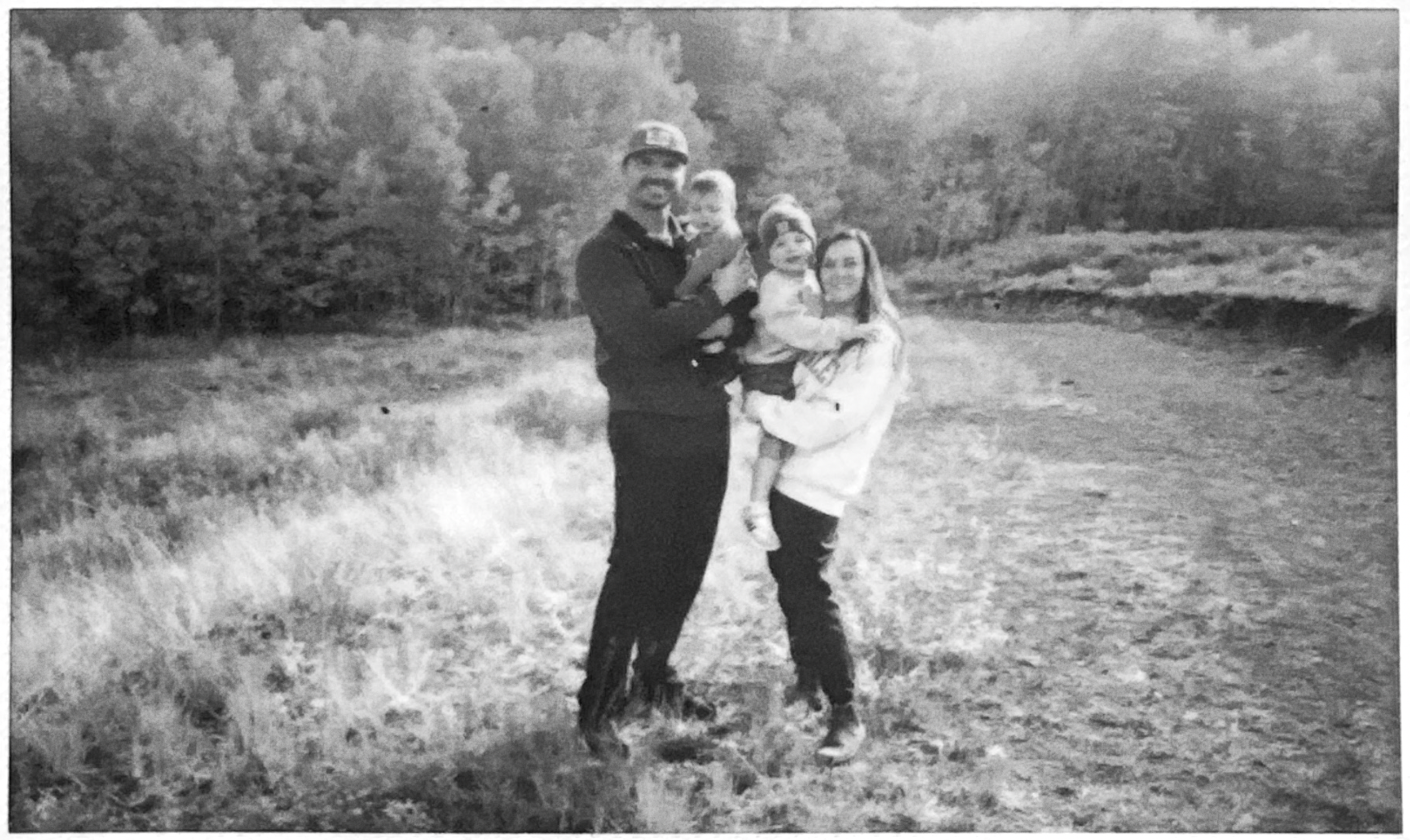 Black and white photo of a family of four standing outdoors on a grassy area with trees in the background. The father is holding a young child, and an adult woman is standing next to them, holding a toddler.
