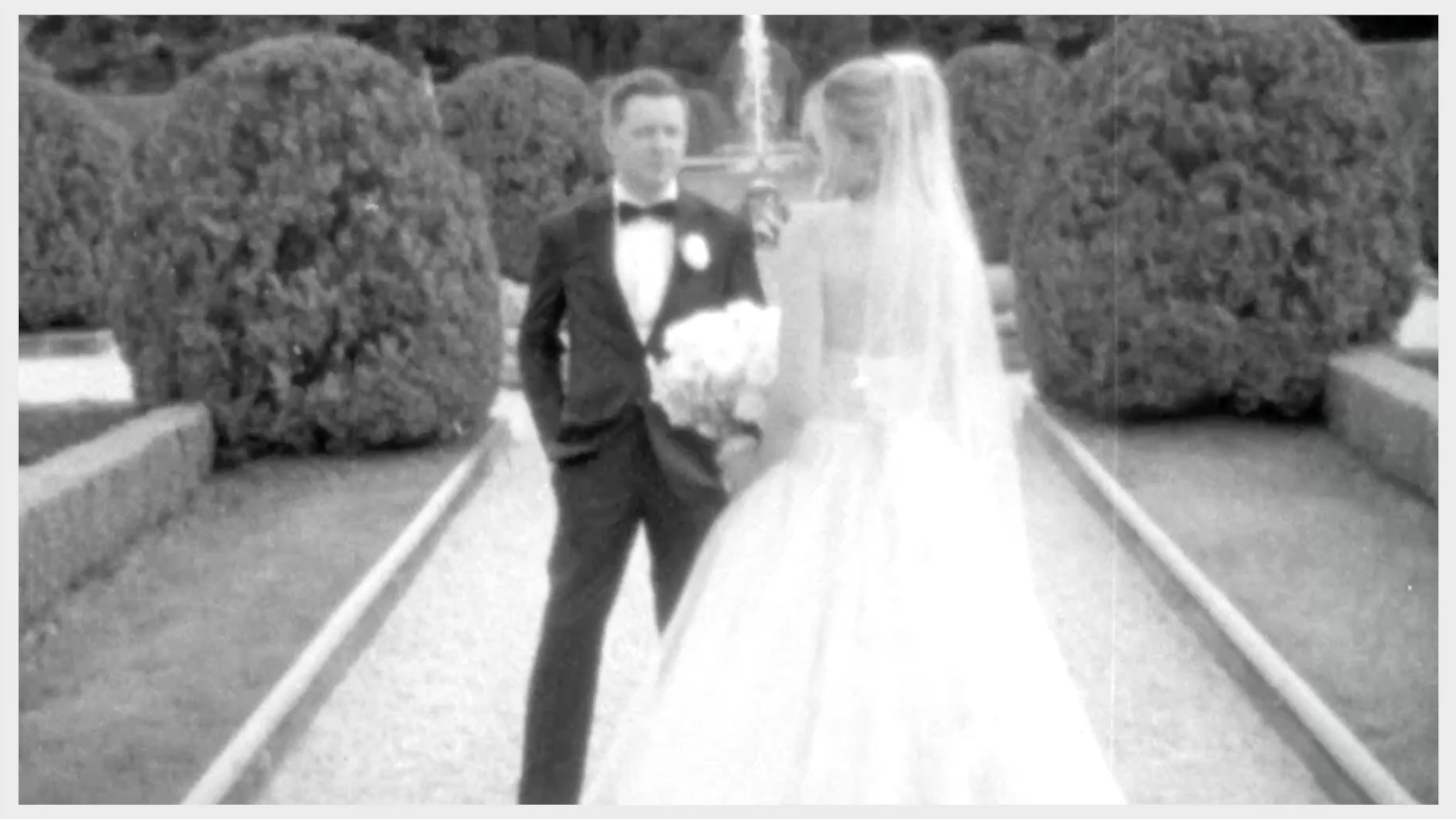 Black and white photo of a bride and groom standing outdoors on a garden pathway. The groom is wearing a tuxedo with a bow tie and holding a bouquet, while the bride is in a wedding gown with a veil.