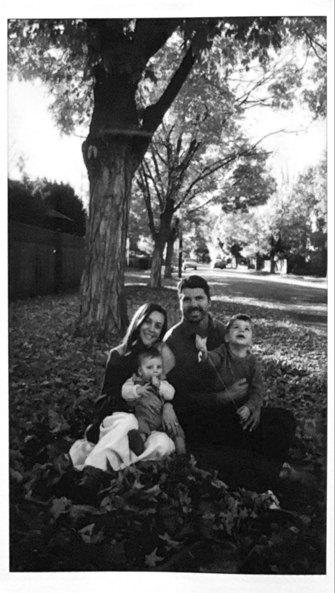 A black and white photo of a family of four sitting on the ground covered with fallen leaves under a large tree in a park or neighborhood. The woman is holding a small child on her lap, the man is sitting next to her with a young boy on his lap. They are all smiling and looking at the camera with trees and a street in the background.