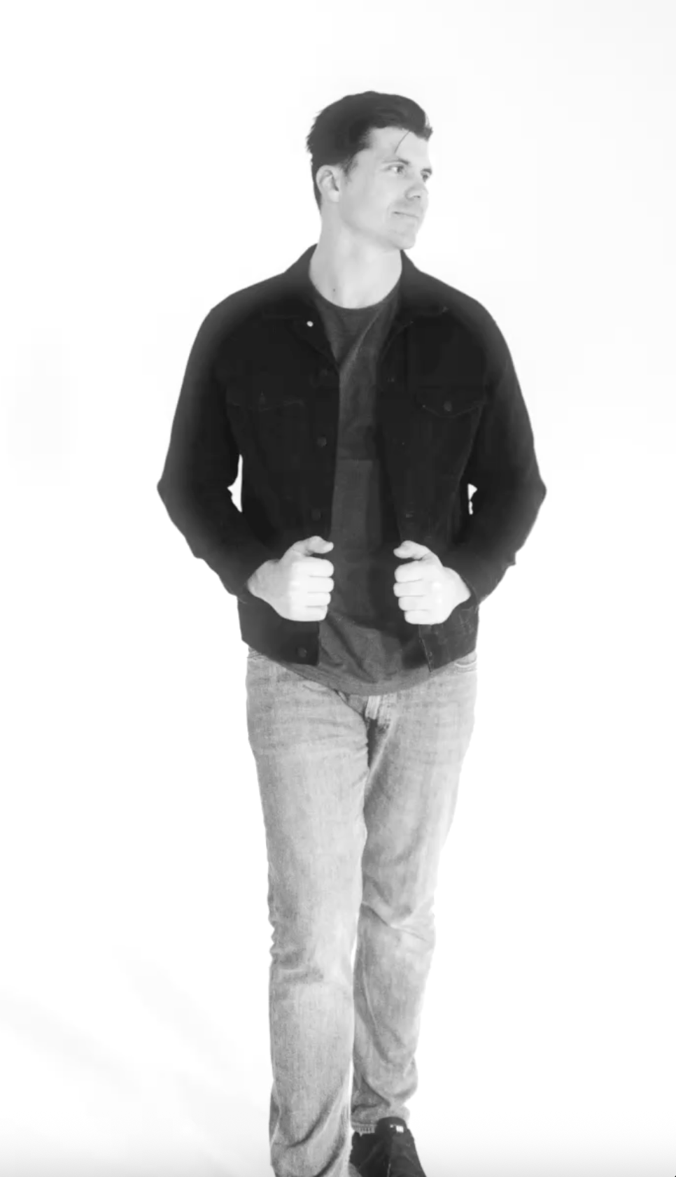 A young man with dark hair, wearing a black denim jacket over a dark shirt, light jeans, and black shoes. He is standing and looking to his right against a plain white background.