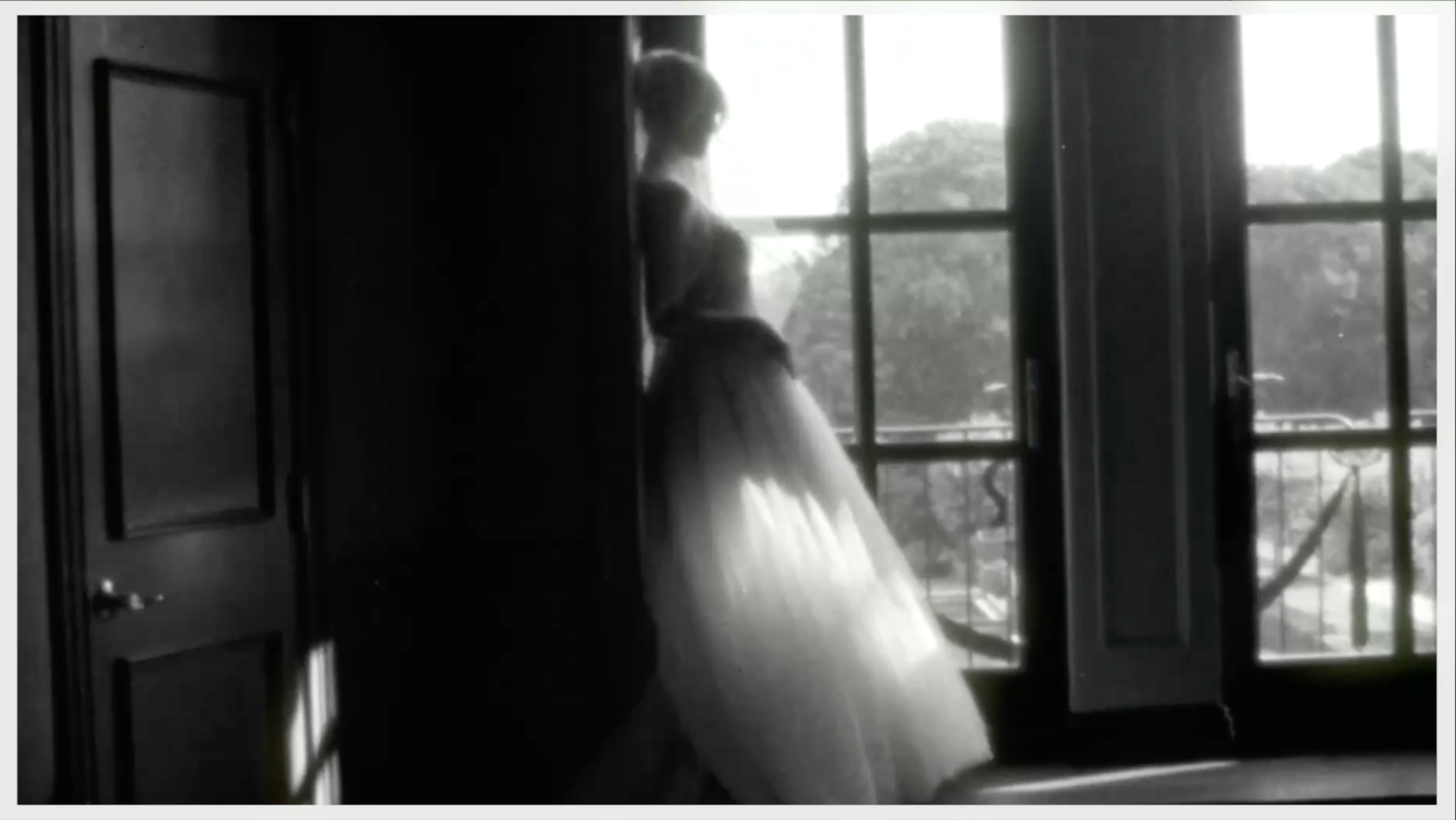 A black and white photo of a girl in a dress sitting by an open window.