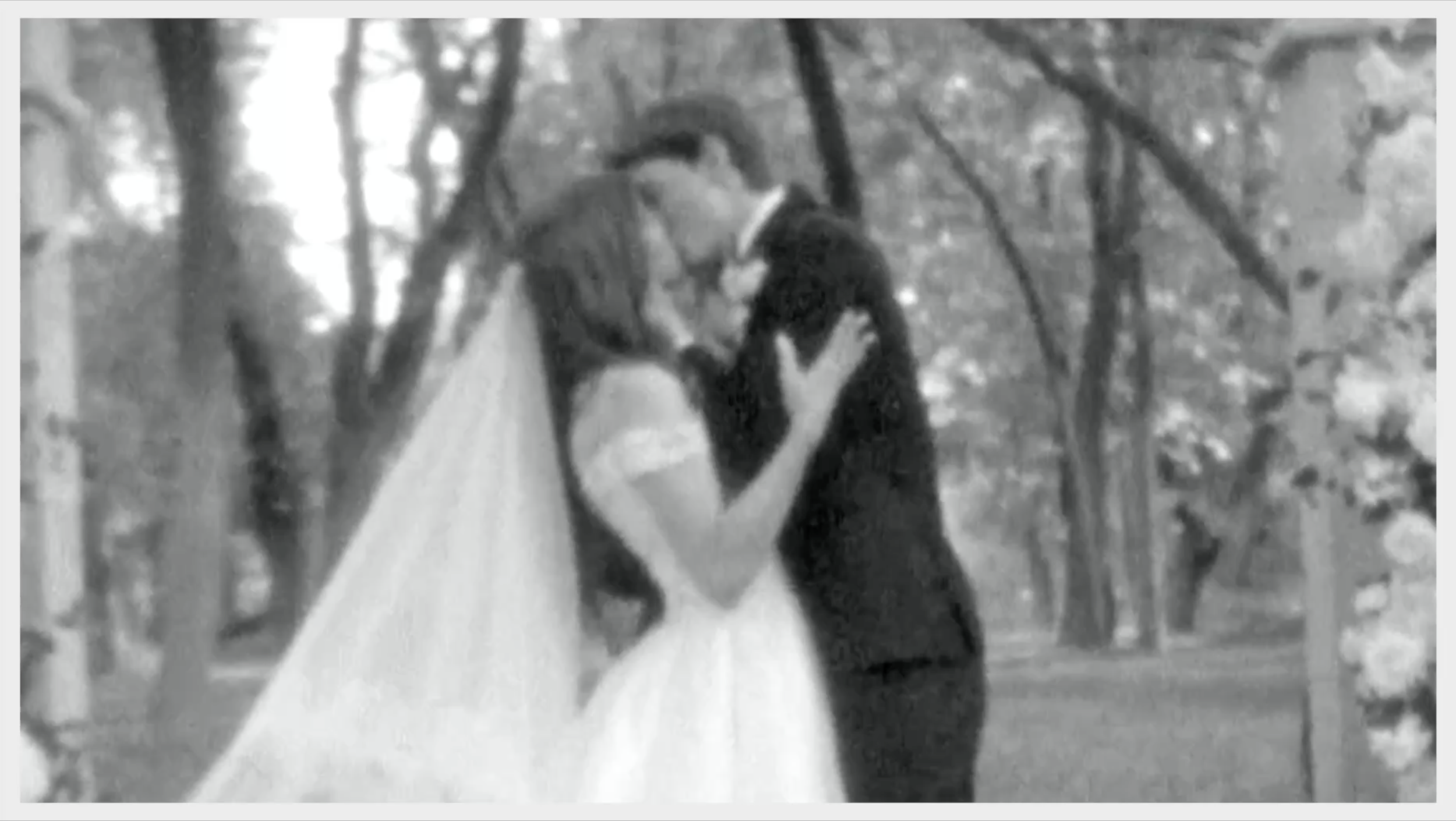 A black-and-white photo of a young couple kissing outdoors in a wooded area, with a girl wearing a white dress and a boy in a dark suit.