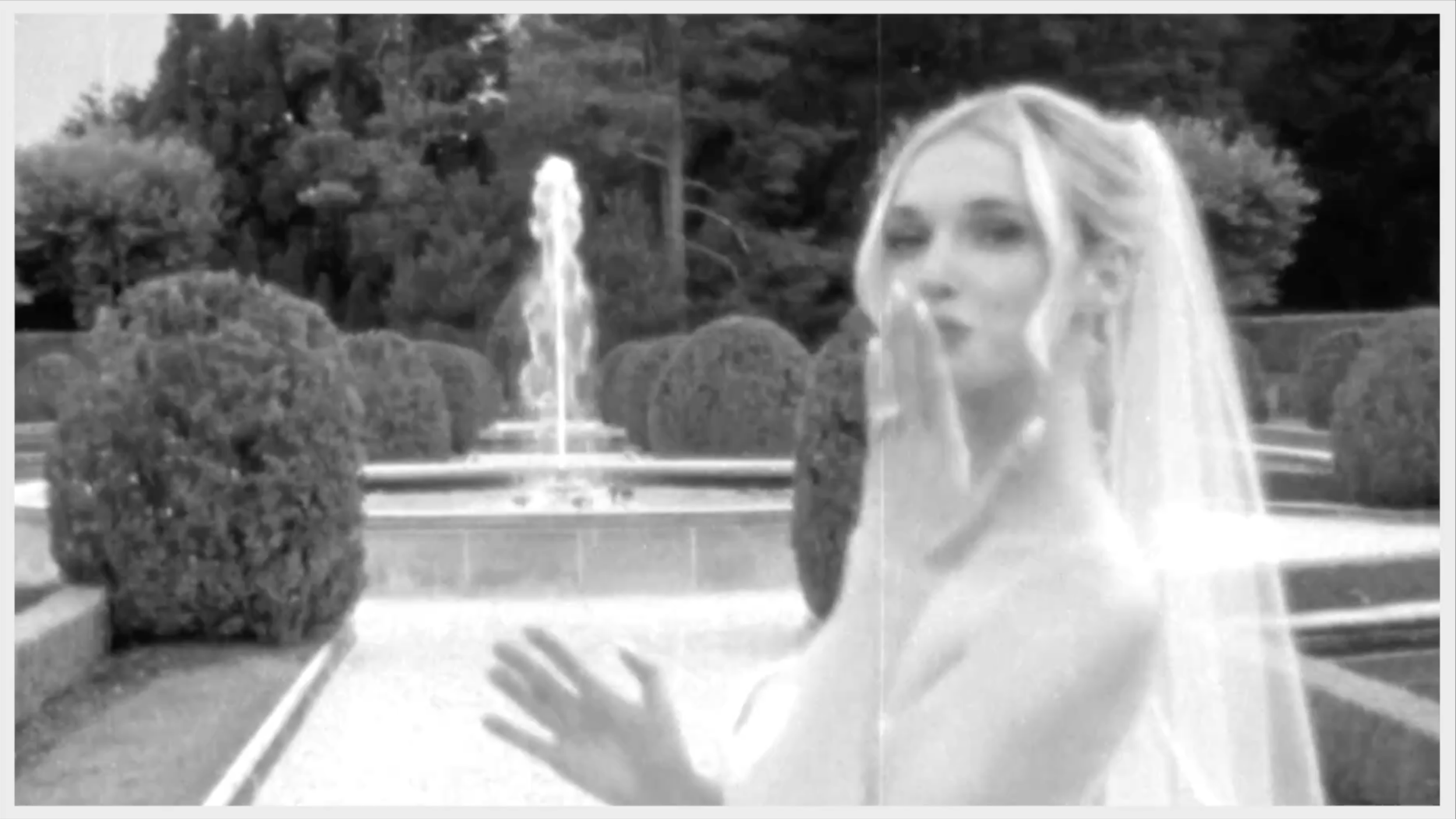 A woman in a wedding dress and veil standing near a water fountain in a garden with shrubs and trees.